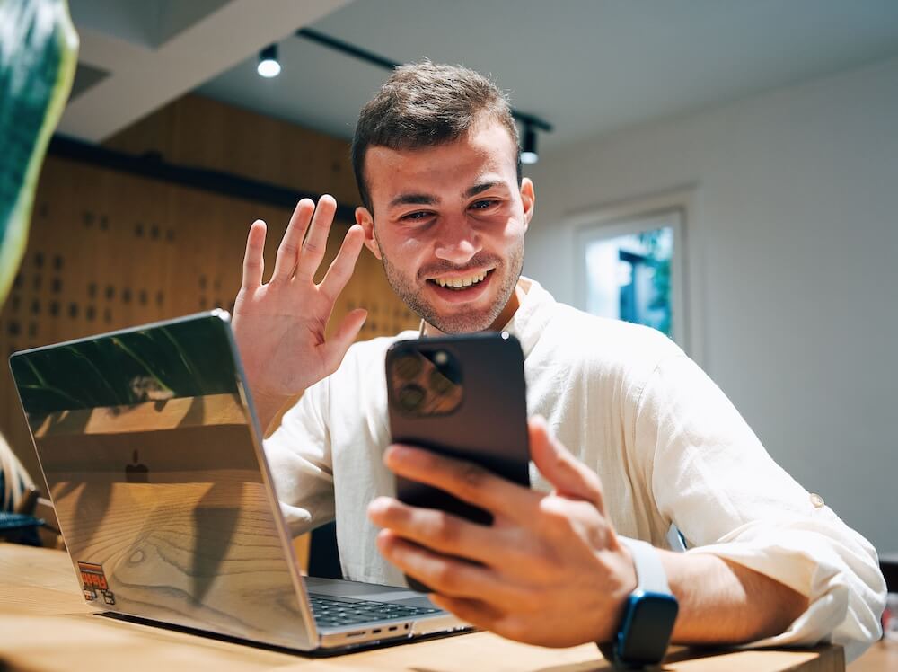 Young man in a white linen shirt grinning and waving at his phone while sitting at a wooden desk with a laptop open | therapeutic coaching online | trauma informed coaching | international trauma therapist | Los Angeles | Seattle | Chicago