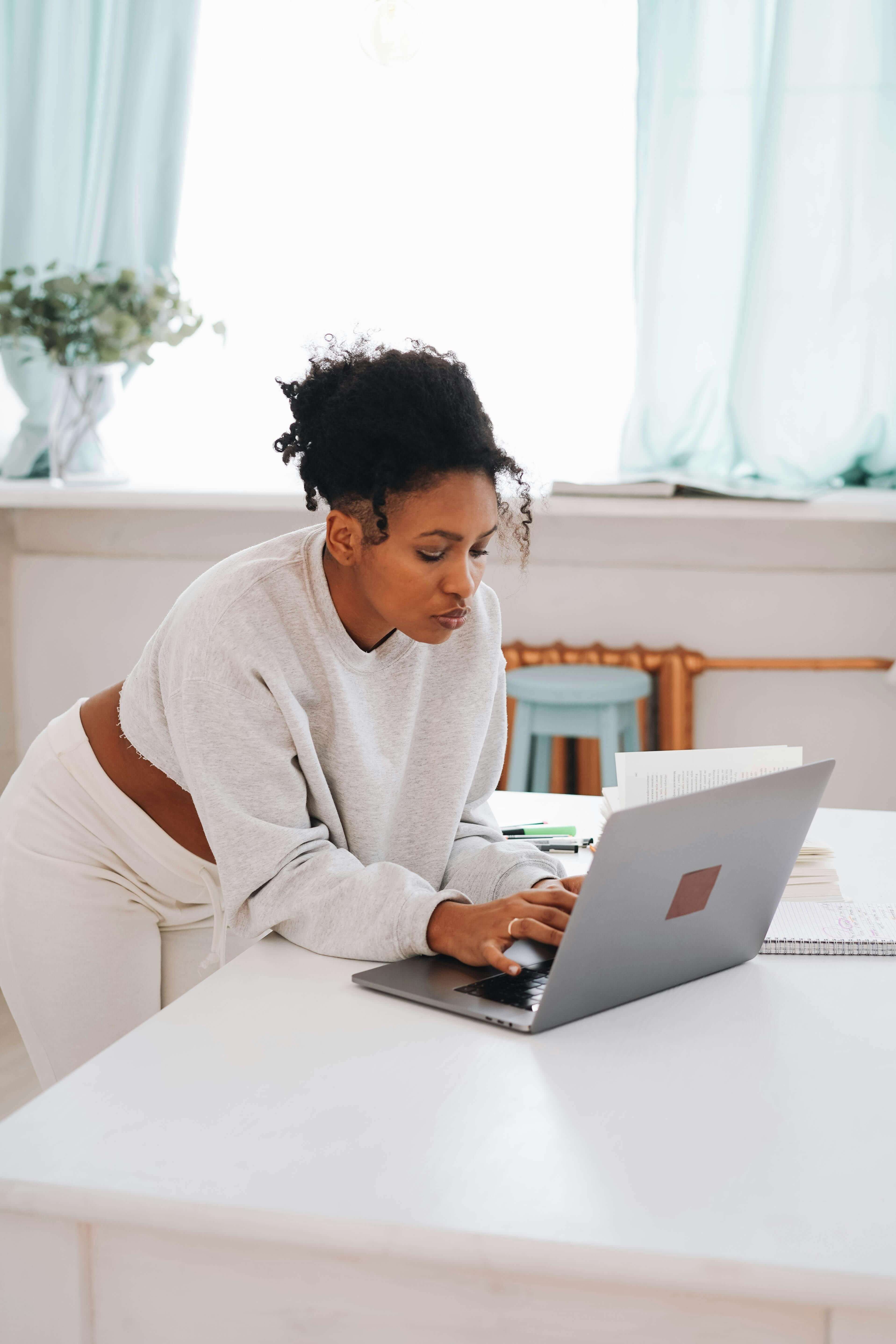 Woman with natural hair in a grey sweatshirt leaning over a white desk working intently on a laptop | international therapist | therapy for expats | why can't I settle down | United States | Canada | Scotland