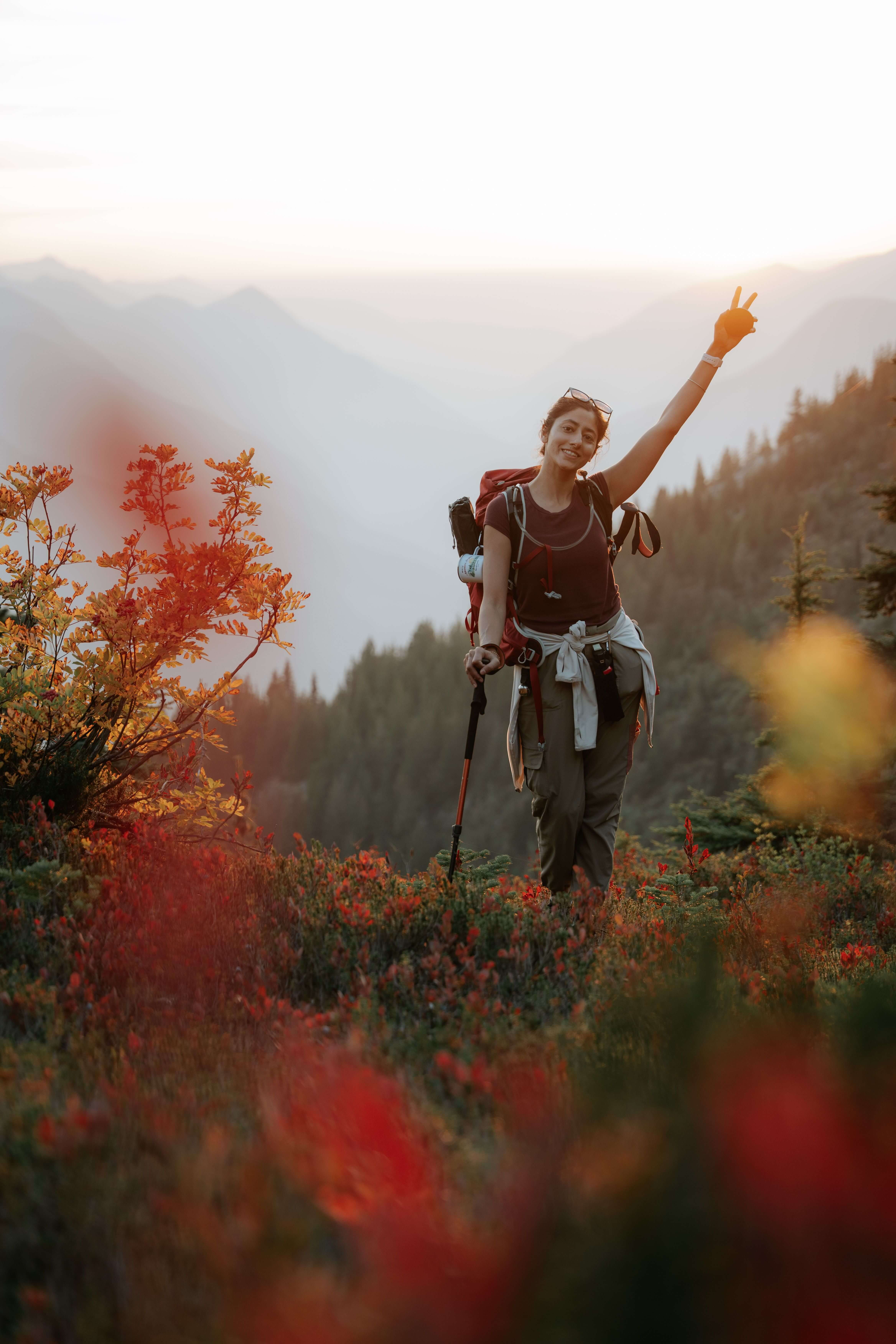 Woman with a large hiking backpack and trekking pole smiling and holding up a peace sign while walking through vibrant red autumn foliage on a mountain trail | therapeutic coaching online | international therapist | parts work training | Seattle | New York City | Chicago