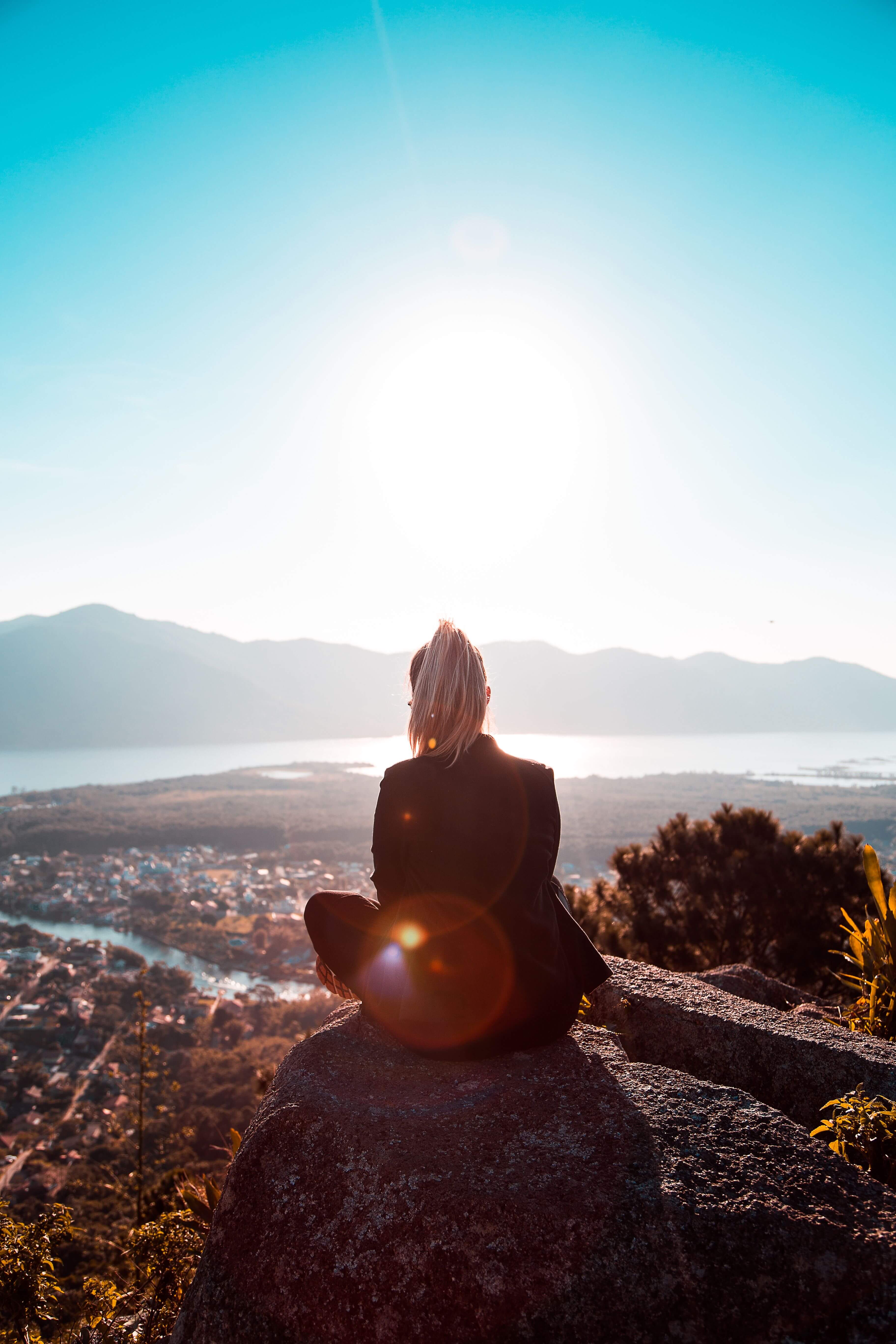 Woman with a blonde ponytail sitting cross legged on a rocky hilltop looking out over a sunlit coastal city and mountains | therapeutic coaching online | energy psychology online | international trauma therapist | Los Angeles | Seattle | New York City