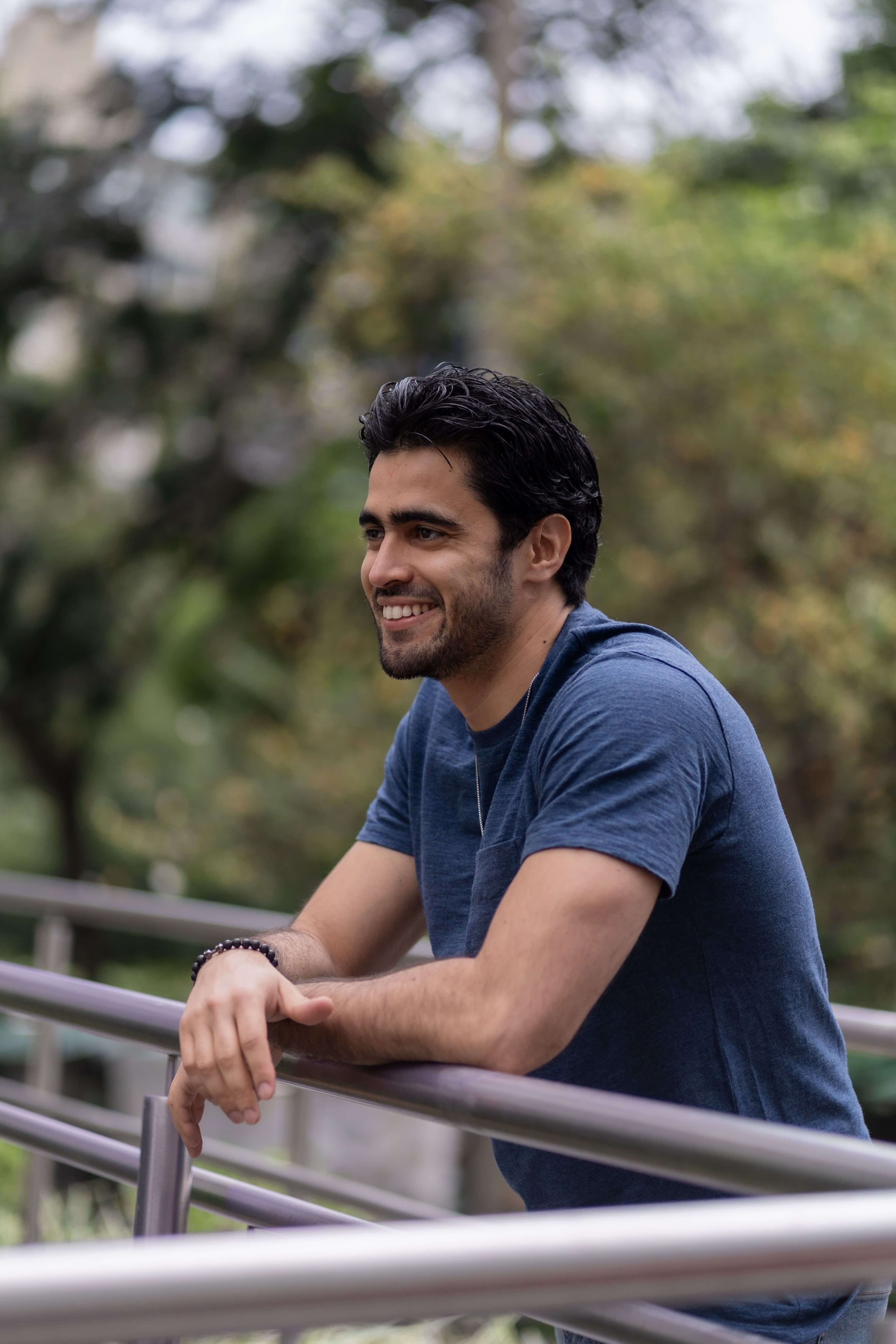 Young man in a blue t-shirt smiling and leaning on a metal railing outdoors surrounded by lush green trees | therapeutic coaching online | international trauma therapist | therapeutic coaching | Seattle | Los Angeles