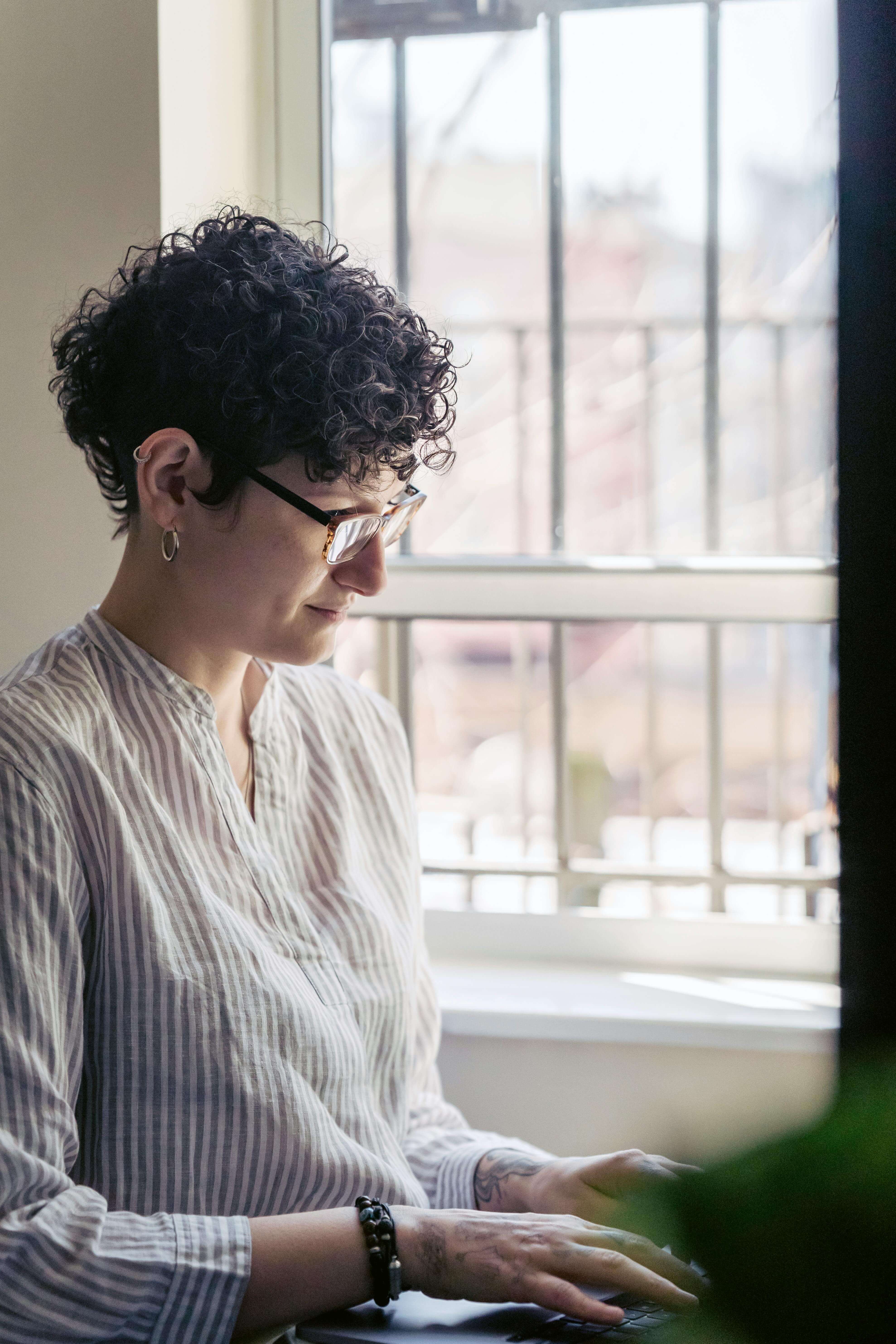 Woman with short curly hair and glasses working at a computer by a bright window in a quiet indoor space | online therapy for expats | international therapy and coaching | therapy across borders | United States | Canada | Scotland