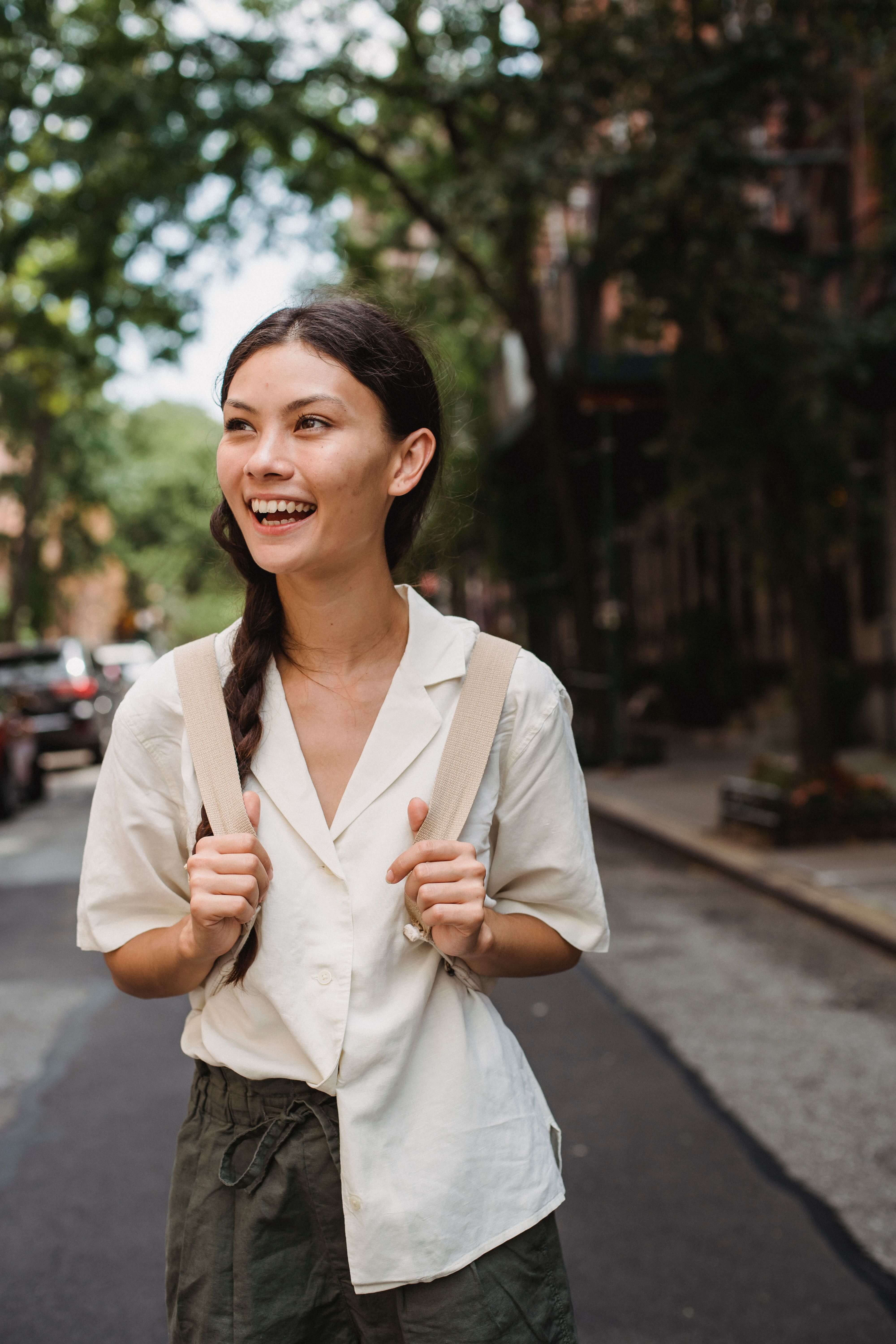 Young woman with a braid and backpack straps smiling and looking up on a tree lined city street | deep connection expat | expat social connection | international therapy and coaching | South Africa | Canada | England