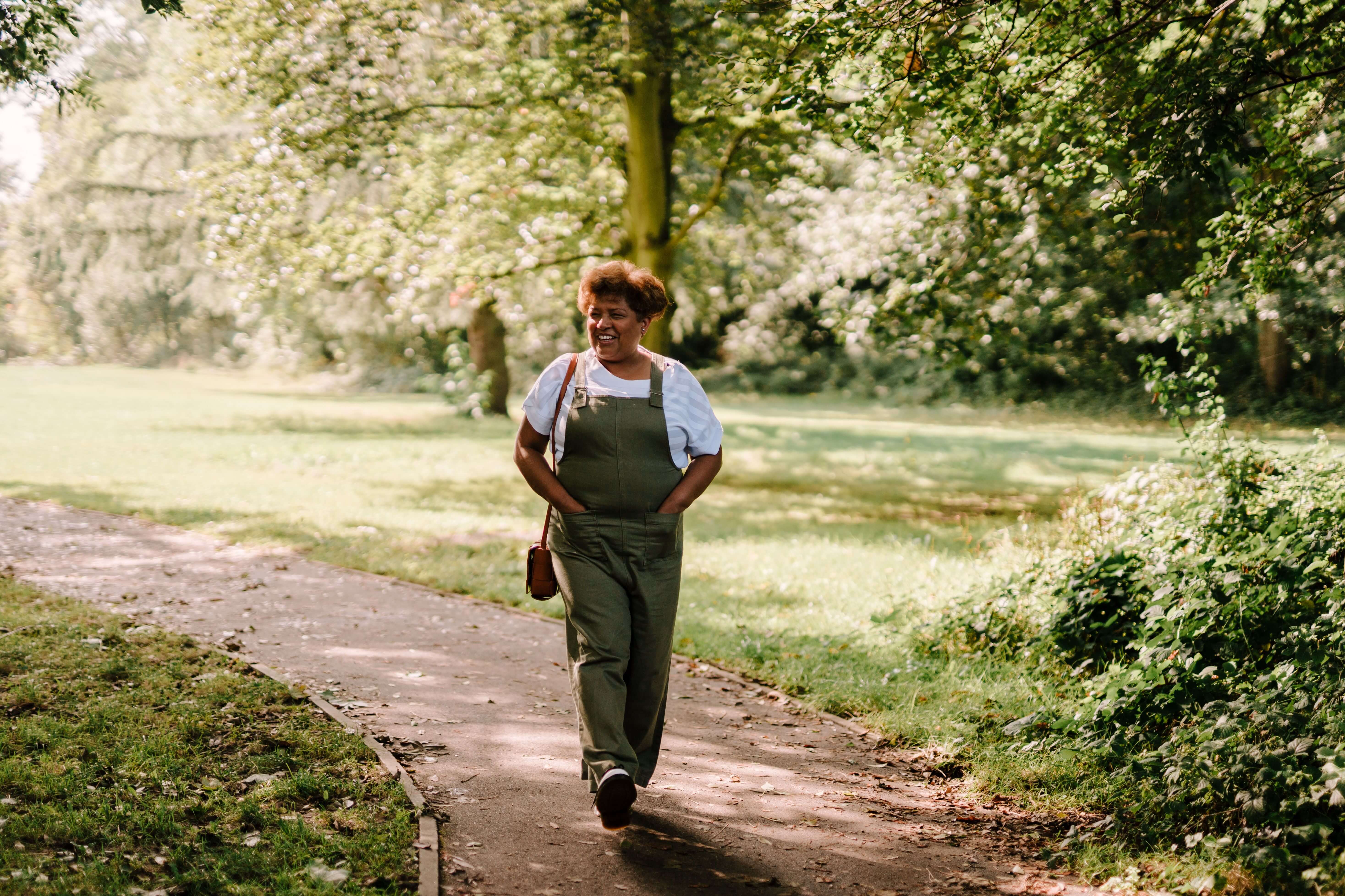 Woman in green overalls smiling and walking along a sunlit park path surrounded by blossoming trees | international therapy and coaching | expat attachment | globally mobile relationships | South Africa | England | Netherlands