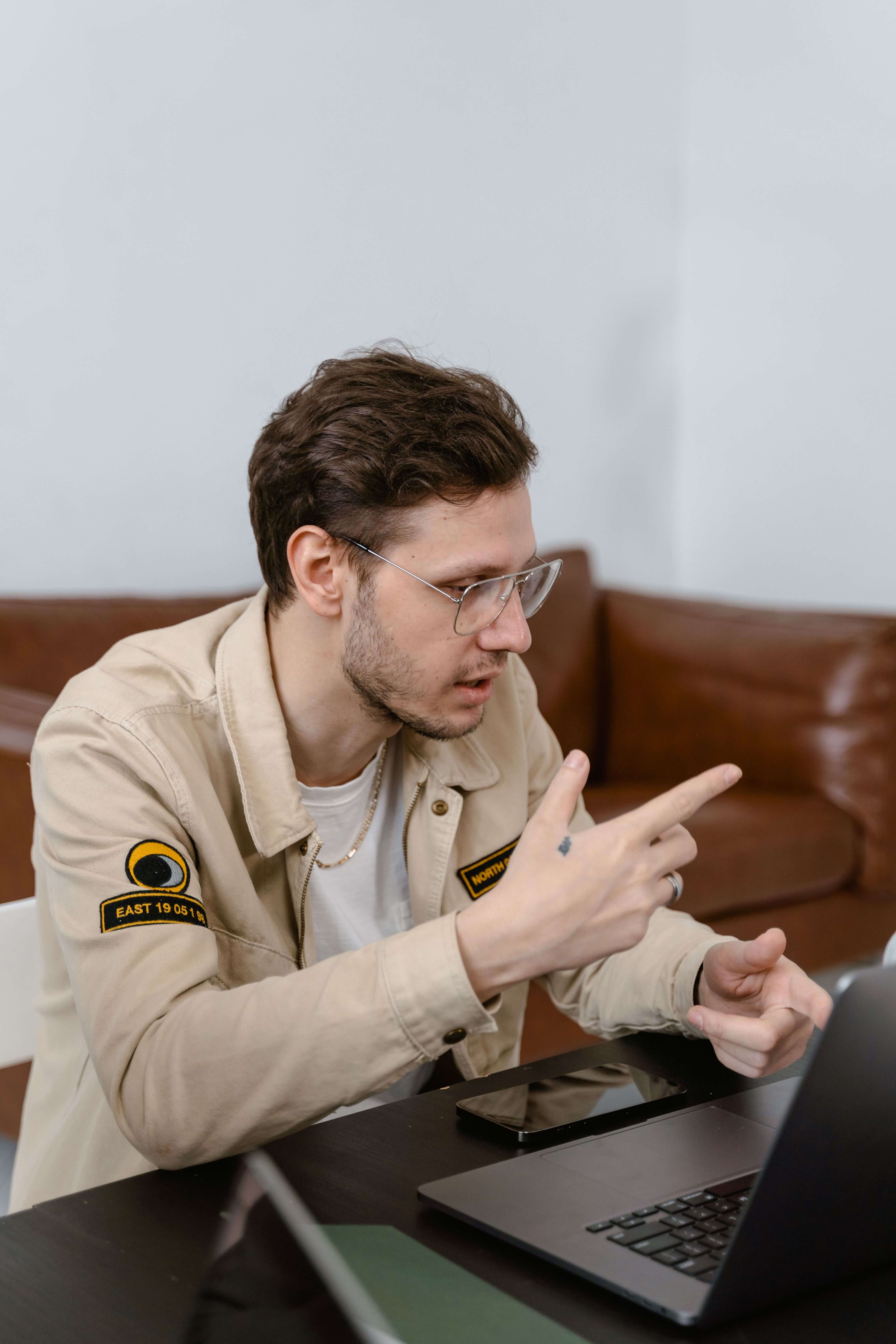 Man with glasses in a beige jacket leaning forward at a desk pointing at a laptop screen with a phone beside him | what is therapeutic coaching | international trauma therapist | trauma informed coaching | Los Angeles | Seattle | Chicago