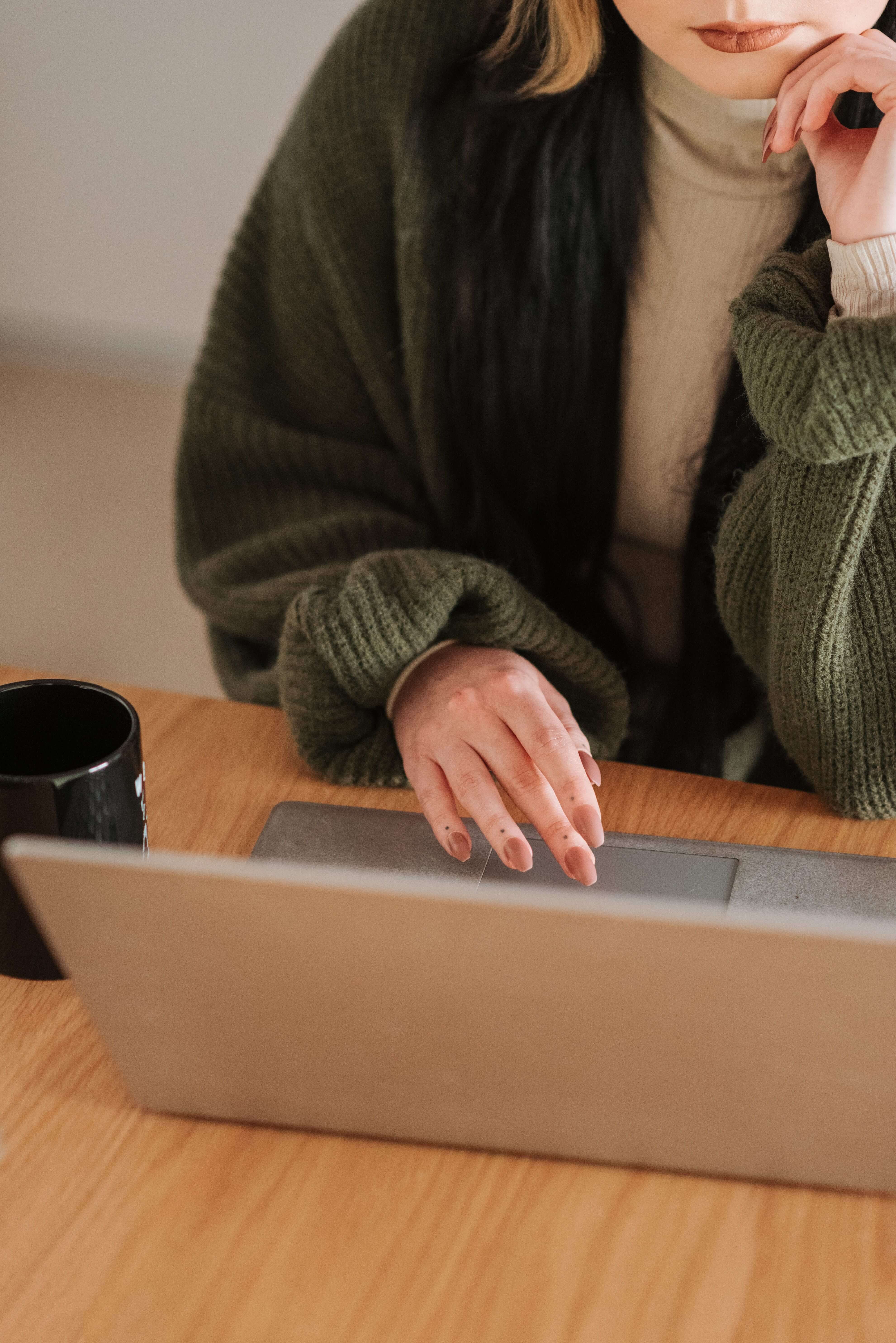 Woman in a green oversized cardigan and cream turtleneck resting her chin on her hand while using a laptop at a wooden desk with a black mug | therapeutic coaching | international trauma therapist | therapeutic coaching online | Los Angeles | Seattle