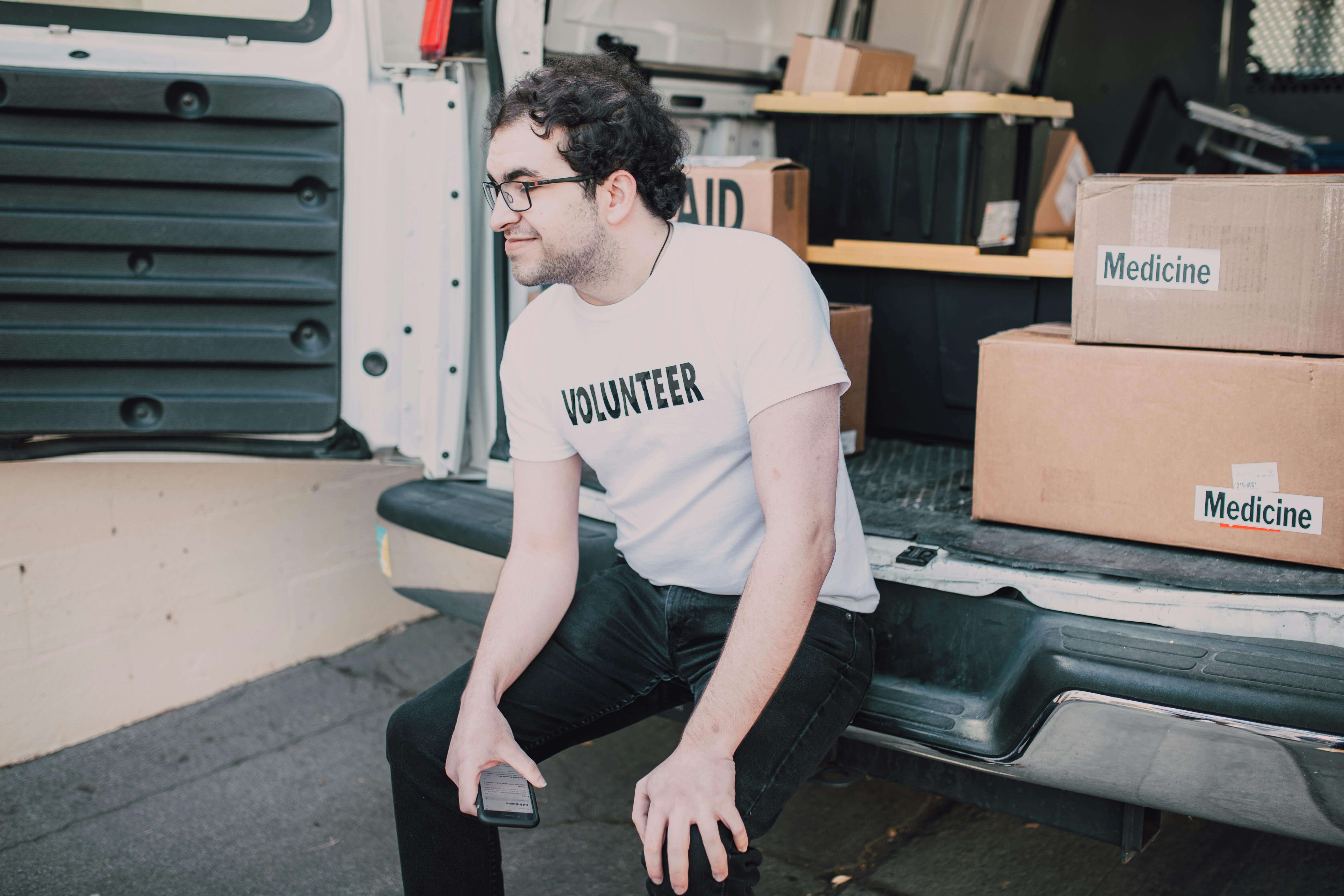 Young man in a volunteer t-shirt sitting on the back of an aid van holding a phone surrounded by medicine boxes | expat sense of self | international therapy and coaching | Germany | Portugal