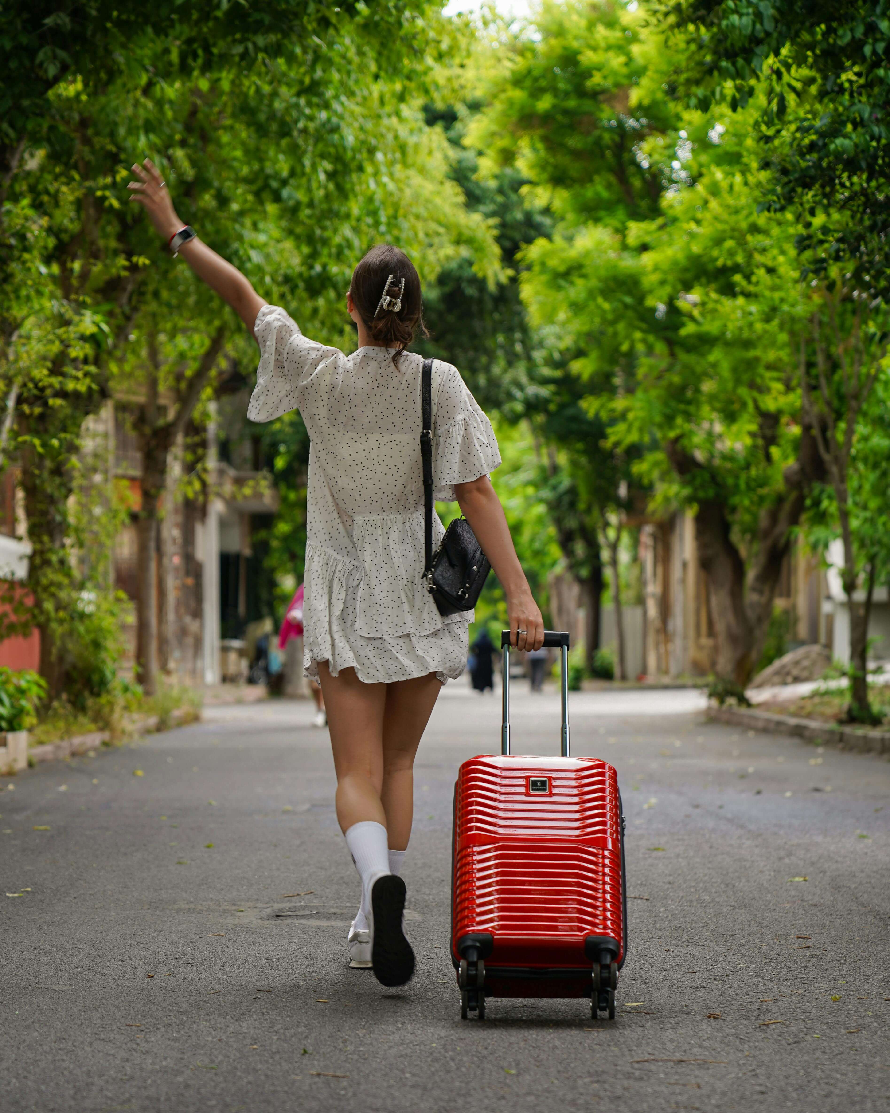 Woman in a white dress walking away down a tree lined street pulling a red suitcase with one hand raised | expat relationship patterns | therapy for expats | love avoidance expat | England | France | Italy