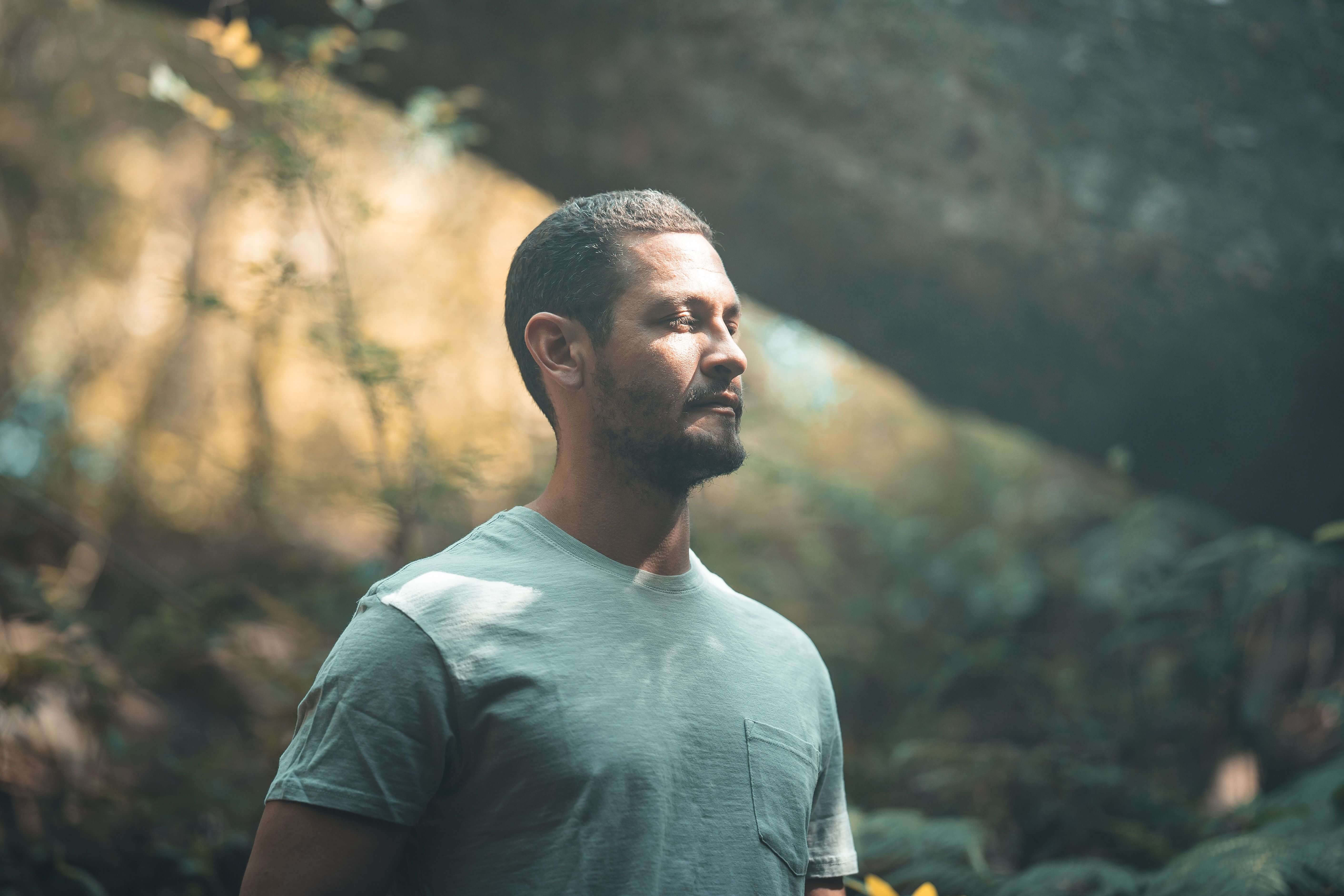 Man in a grey t-shirt standing in a sunlit forest with light streaming through trees looking pensively to the side | energy psychology online | tapping therapy online | international trauma therapist | Chicago | New York City | Seattle