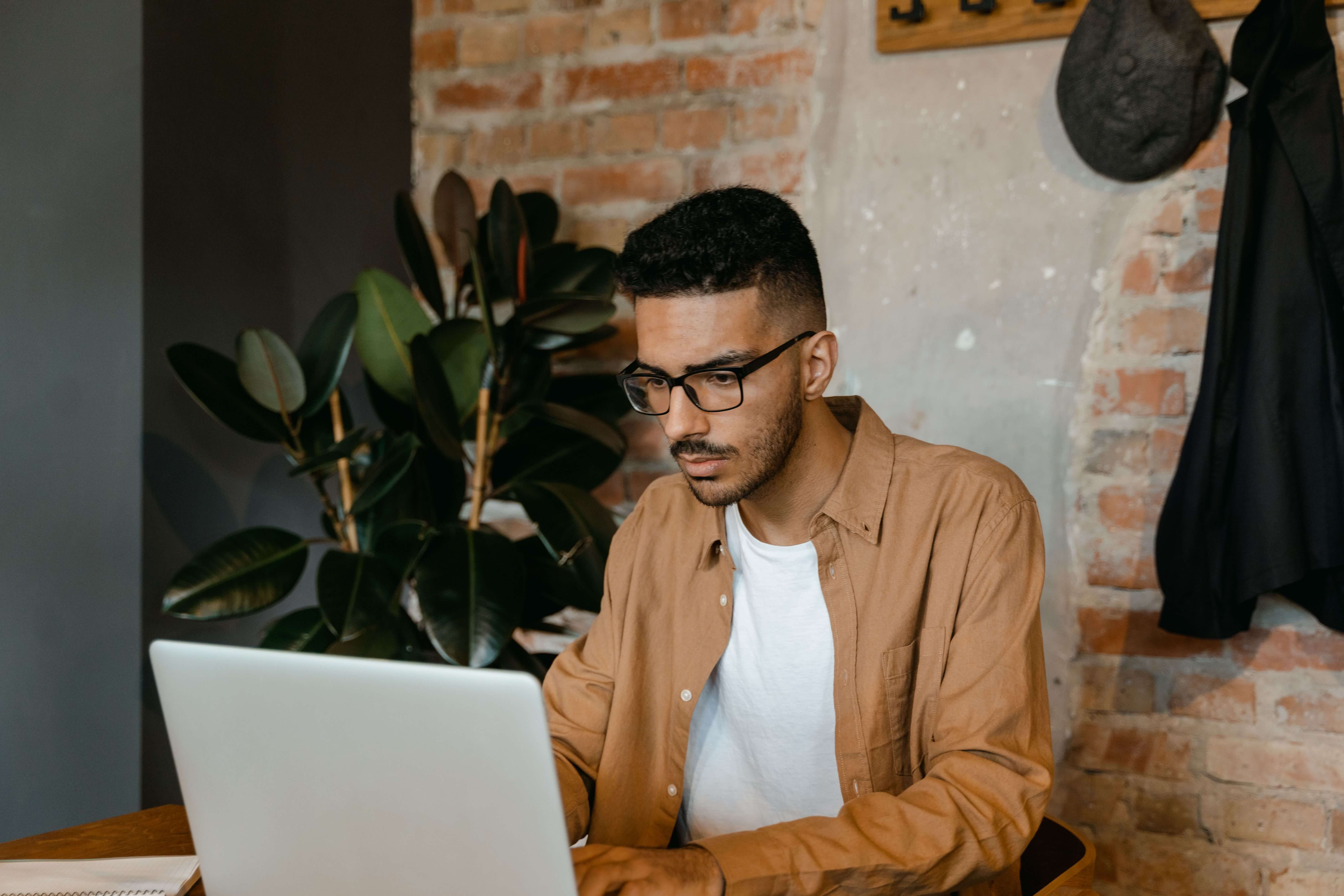 Man with glasses and a brown shirt sitting at a wooden desk working intently on a laptop beside a large leafy plant | EMDR session online | international trauma therapist | virtual EMDR | United States | France | Italy
