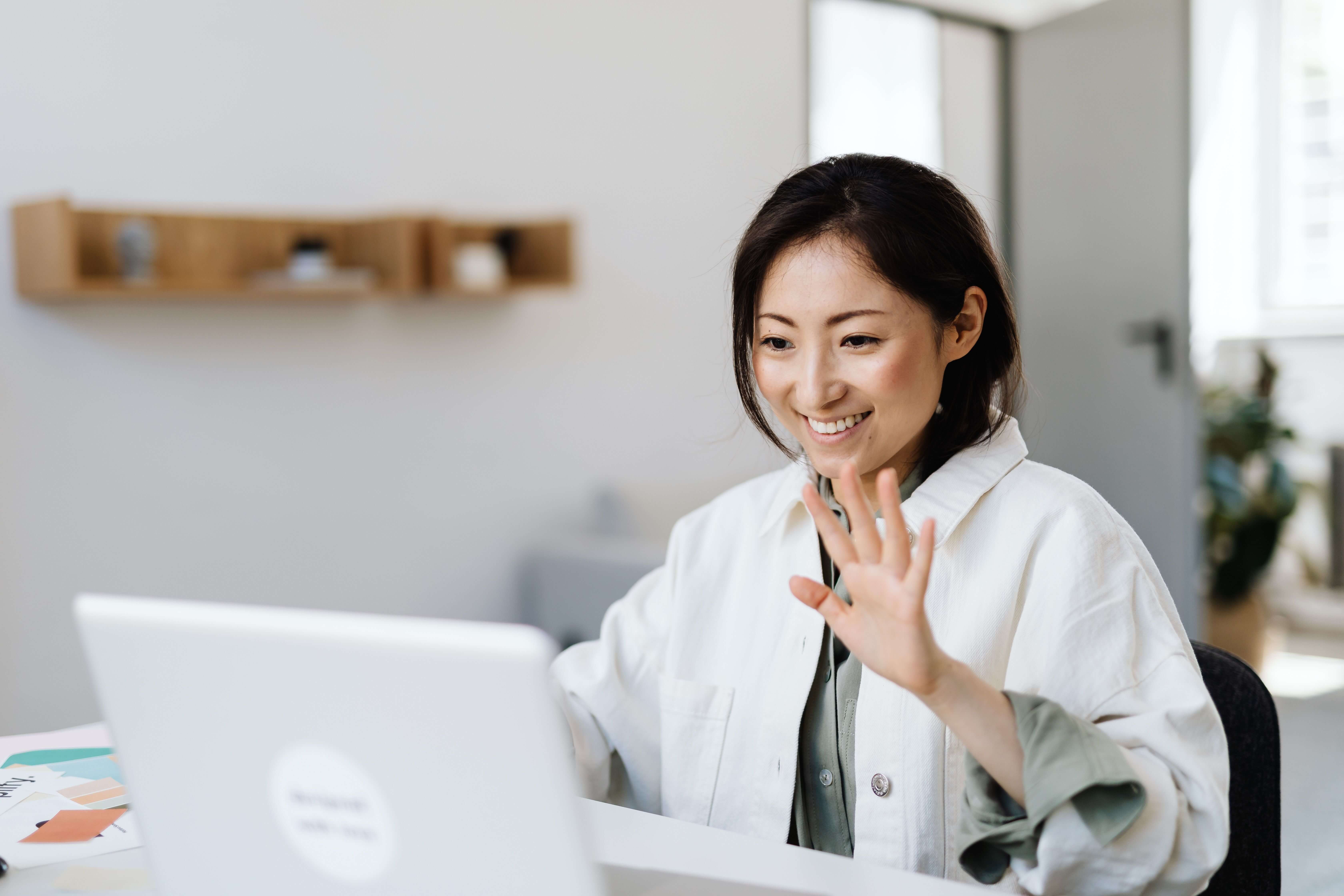 Woman in a white jacket smiling and waving at a laptop screen in a bright minimalist home office with plants in the background | therapeutic coaching online | international trauma therapist | tapping therapy online | Los Angeles | Seattle | New York City