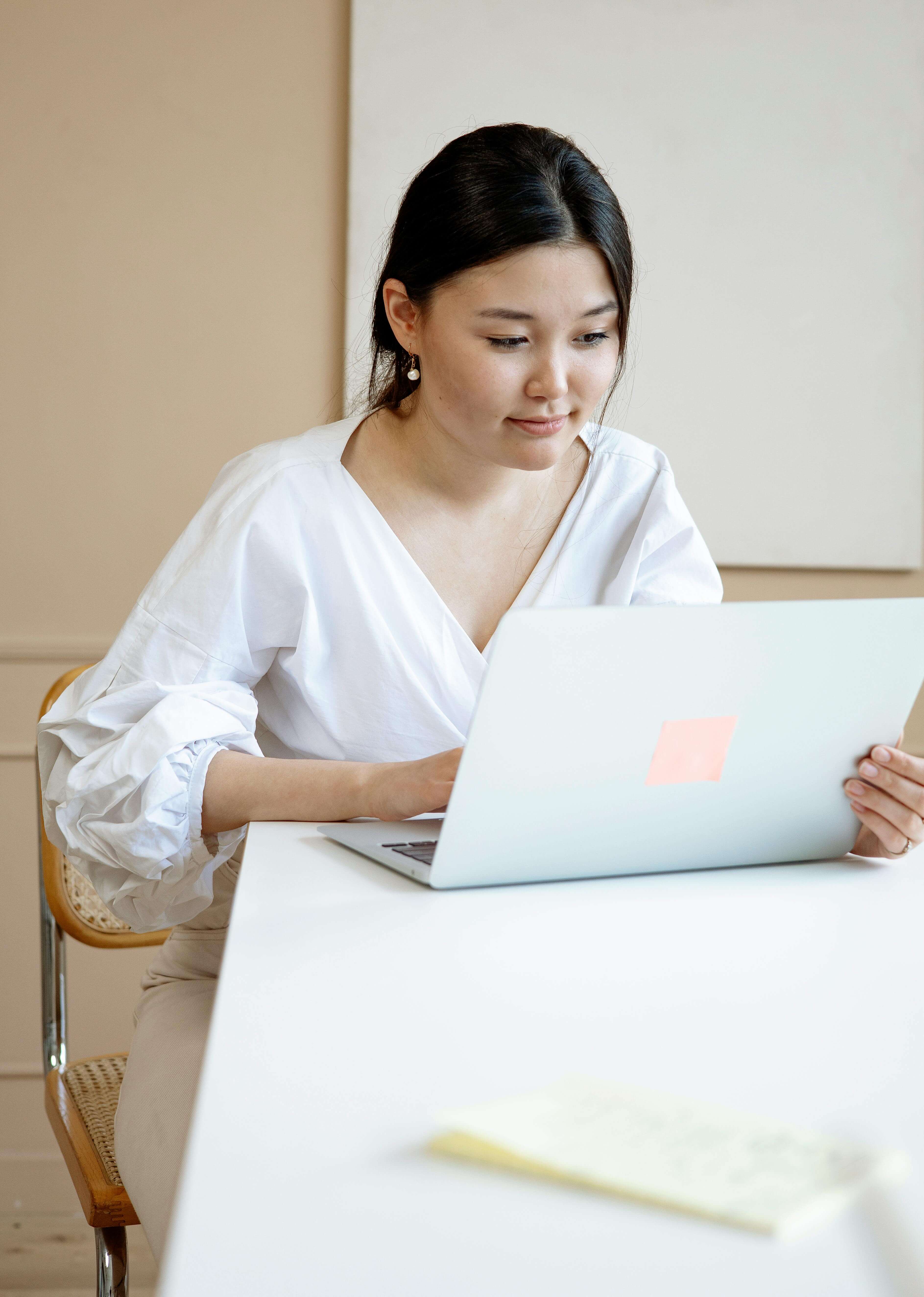 Woman in a white blouse sitting at a white desk working on a laptop during an online therapy session | international therapist | international therapy and coaching | expat nervous system | United States | South Africa | Germany