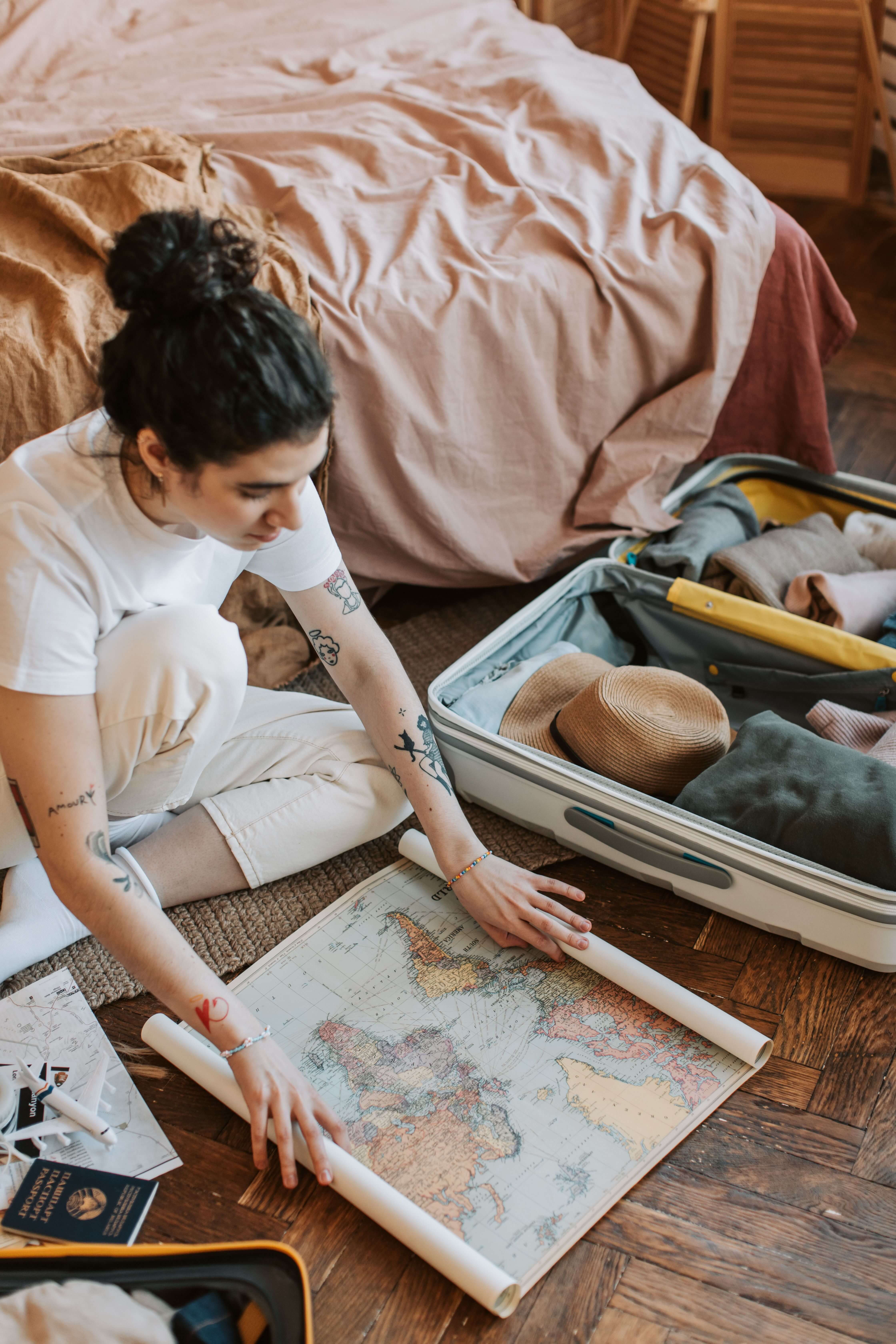Woman with tattoos sitting on the floor beside an open suitcase looking at a world map with a passport nearby | expat love avoidance | internationally mobile relationships | therapy for expats | England | Netherlands | France