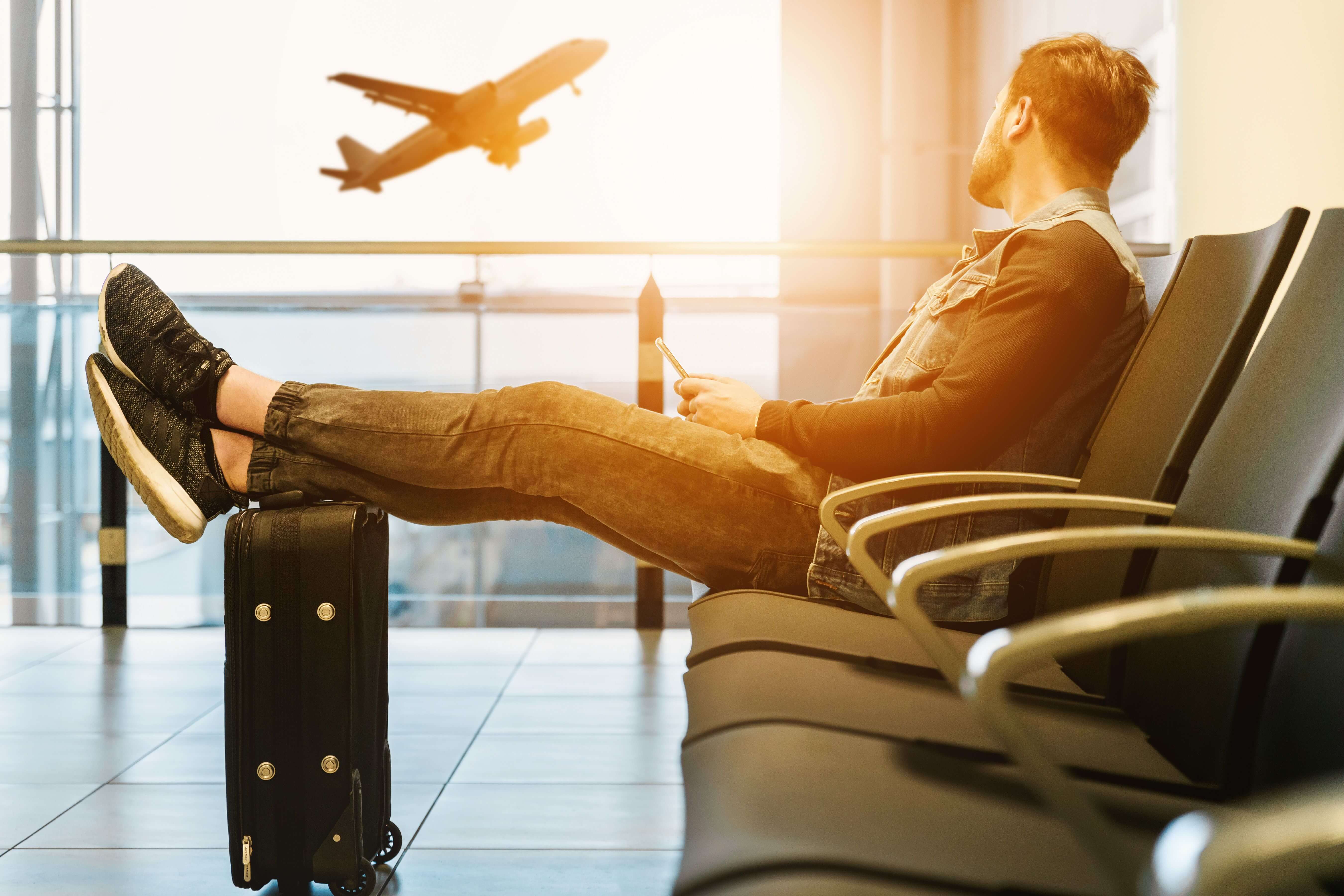 Man sitting in an airport departure lounge with feet resting on a suitcase watching a plane take off through the window | why do I keep moving | running away abroad | fear of settling down expat | Germany | Canada | Italy