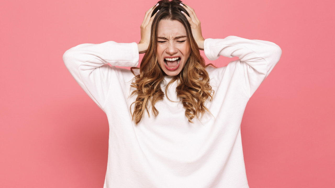 A candid portrait of a visibly overwhelmed and stressed parent, with messy hair holding her head with a weary expression against a solid pink background 