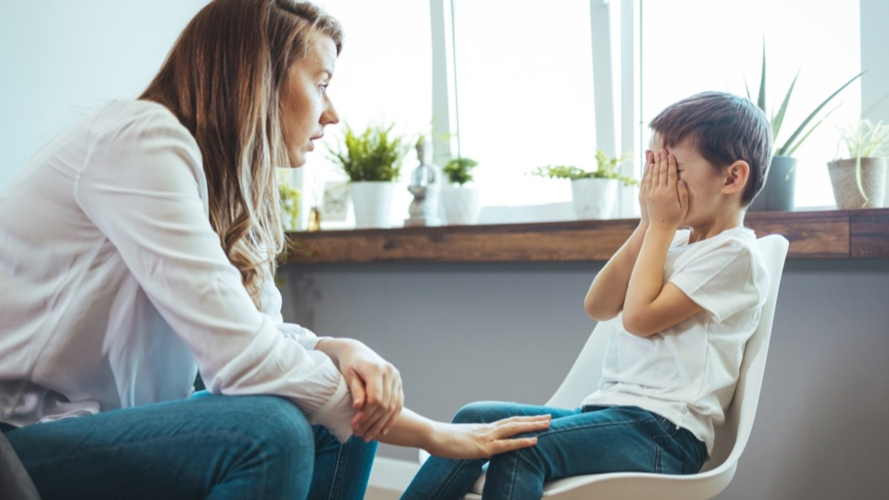 A young boy is upset holding his hands over his eyes, while receiving comfort from a supportive adult sitting near him.