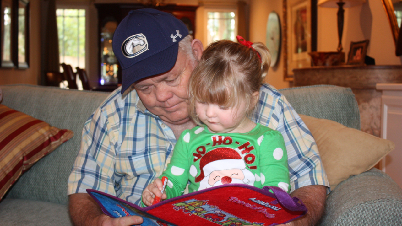 A young girl in a holiday shirt sitting on the lap of her grandfather, looking at a book together