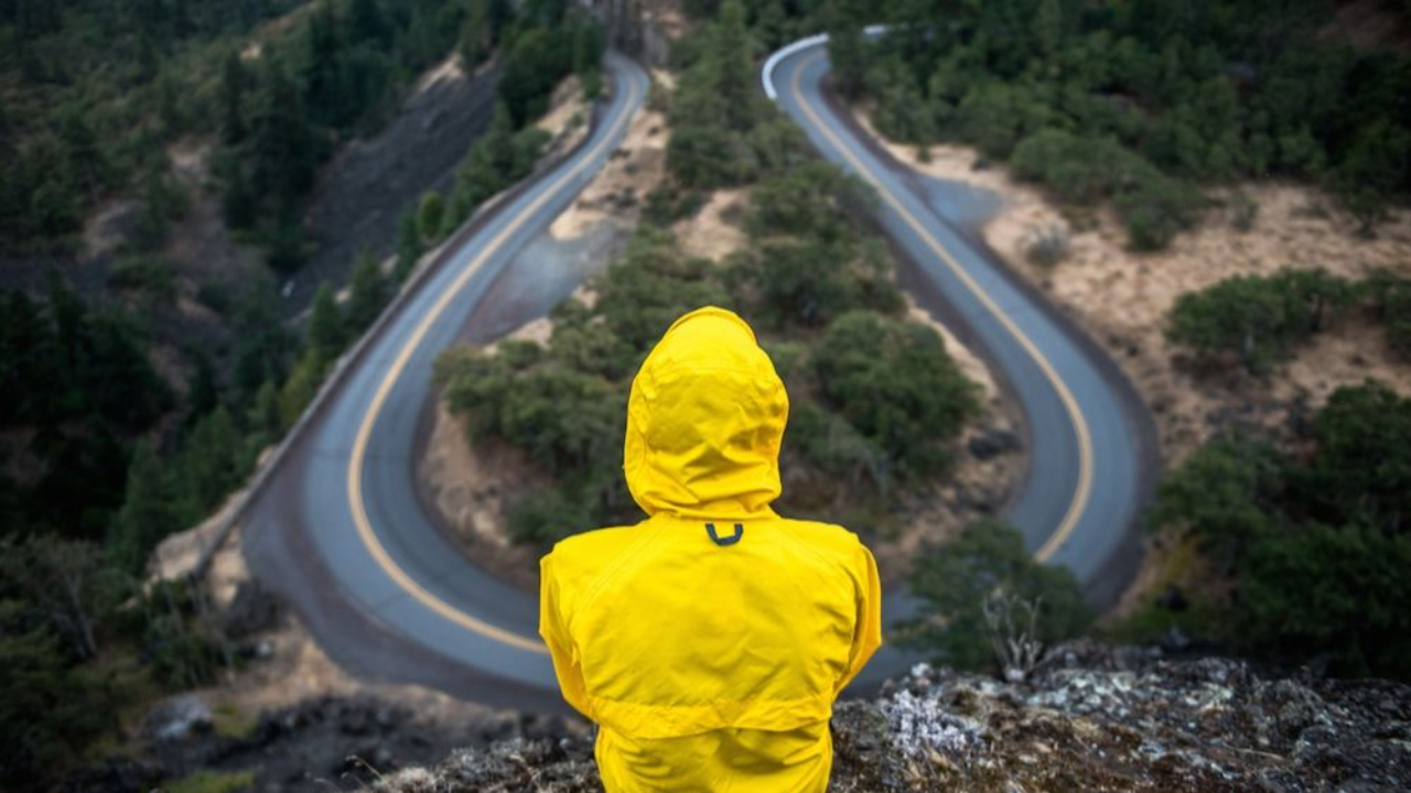 Person in a yellow jacket sitting on a cliff, overlooking a winding road surrounded by trees and hills."