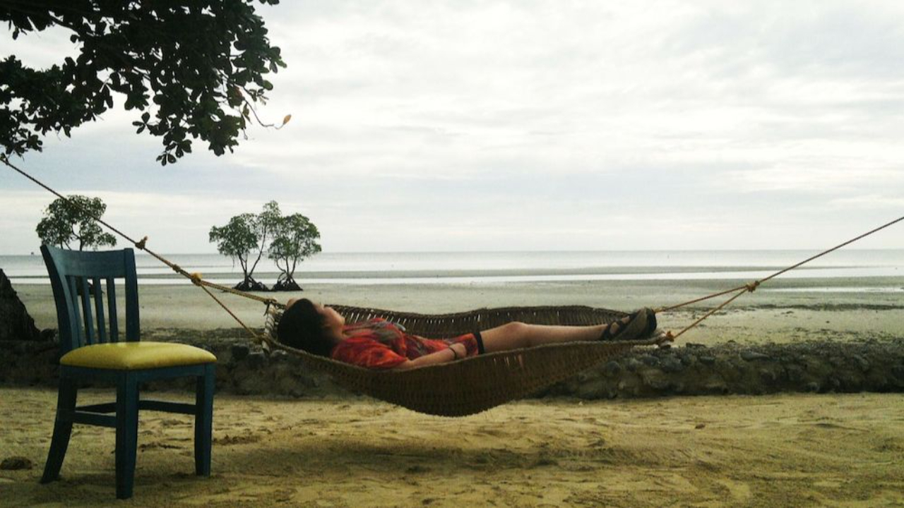 Person relaxing in a hammock on a sandy beach, with a calm ocean view and trees in the background.