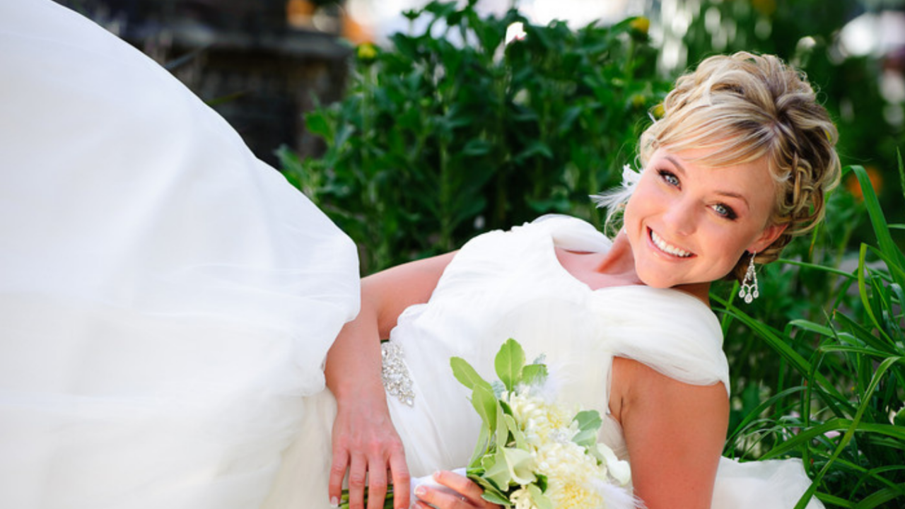Smiling LDS bride in modest white wedding gown holding bouquet, reclining outdoors with soft greenery background
