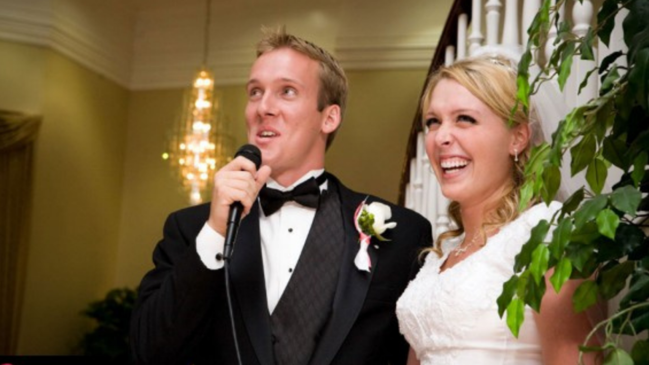 Bride smiling during wedding reception while groom gives a toast, surrounded by guests in a warmly lit LDS reception setting.
