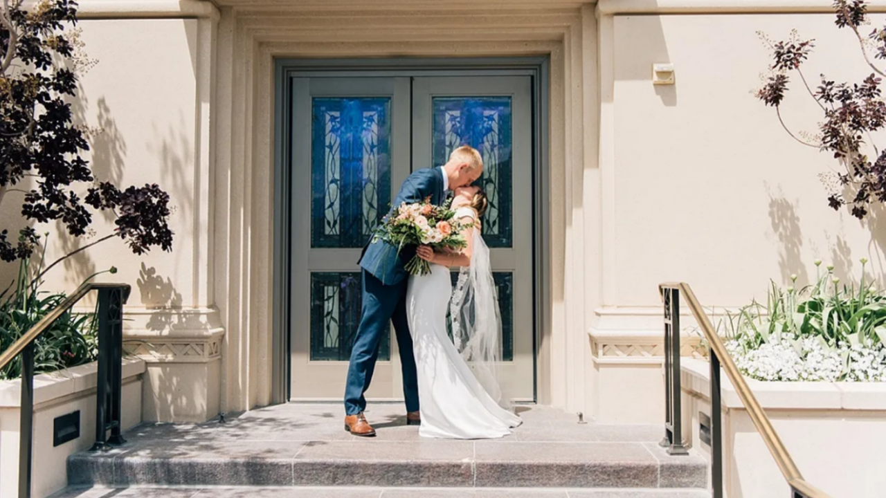 LDS couple kissing outside the Payson Utah Temple after sealing, holding a summer wedding bouquet before their rustic Linden Barn reception.