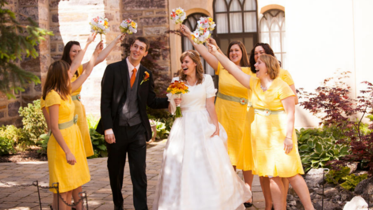 Bride and groom smile joyfully under a bouquet arch formed by bridesmaids in cheerful yellow dresses during a bright summer day at the Logan Utah Temple.