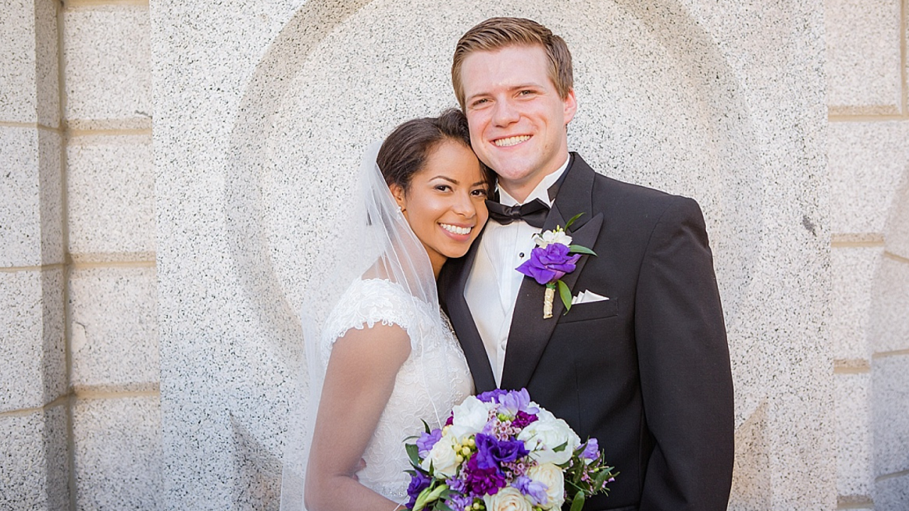 Bride and groom smiling in front of the Salt Lake Temple’s stone archway, holding a purple and white bouquet — image courtesy of Terra Cooper Photography, Church of Jesus Christ Wedding Planning.