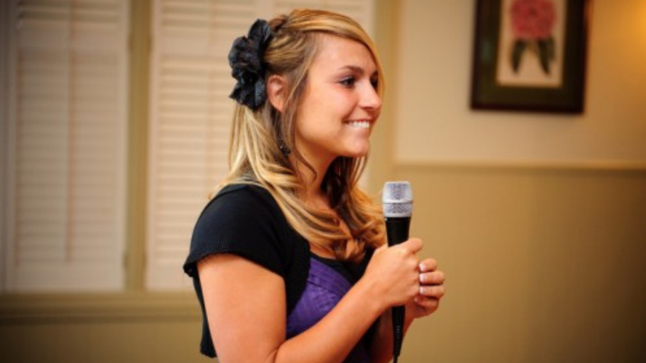 Young woman giving a wedding speech while holding a microphone, smiling warmly during a reception toast.