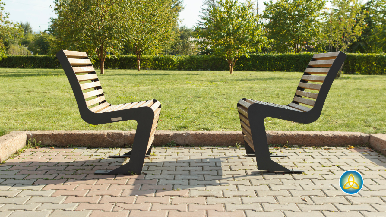 Two empty chairs face each other on a sunlit stone patio surrounded by green space, symbolizing the open, equal relationship between a male therapist and the men he works with