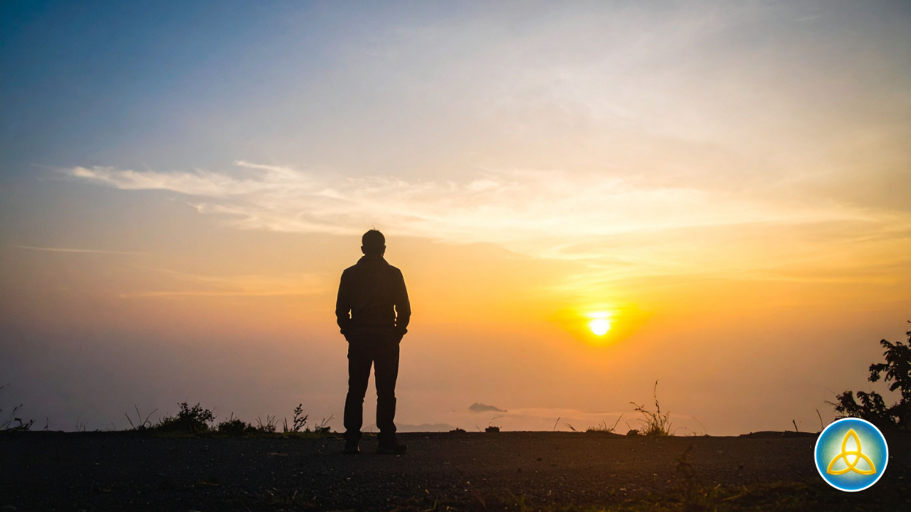 Silhouette of a person standing on solid ground at the edge of a wide landscape, facing a golden sunrise on the horizon