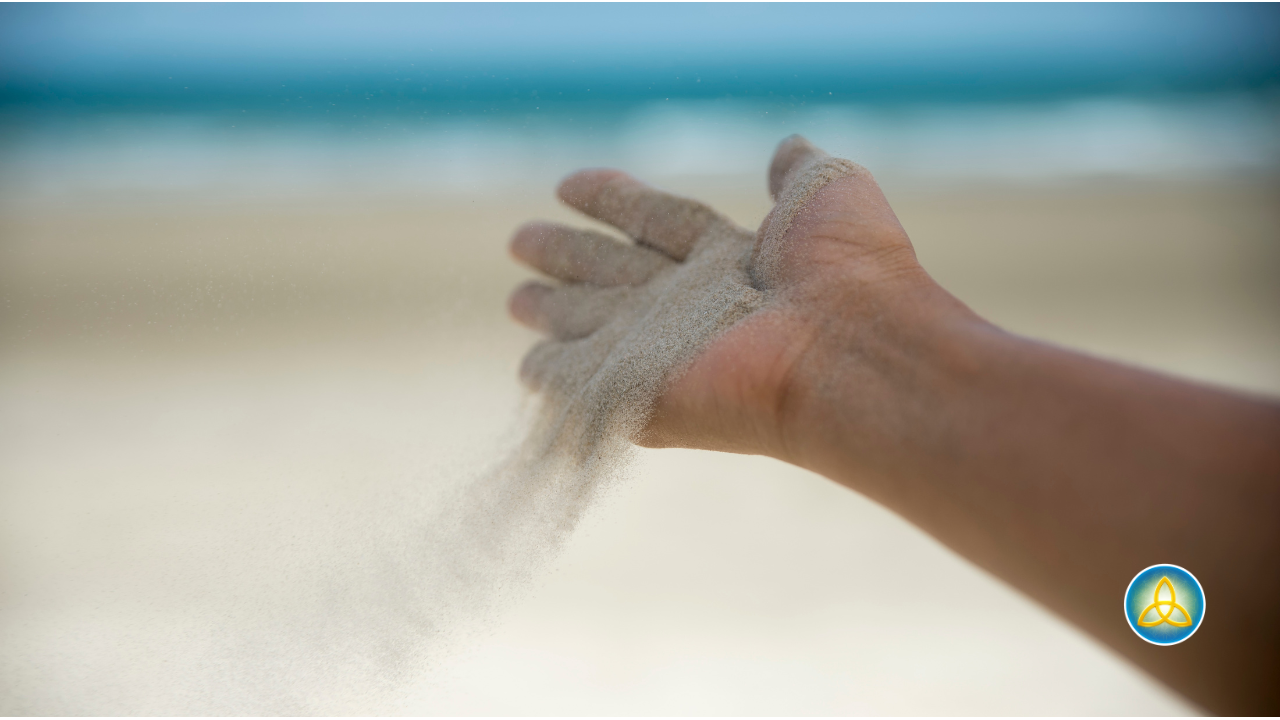 An open hand releasing sand into the wind on a beach, illustrating the practice of letting go of excess potential and the grip of forced effort in practical metaphysics
