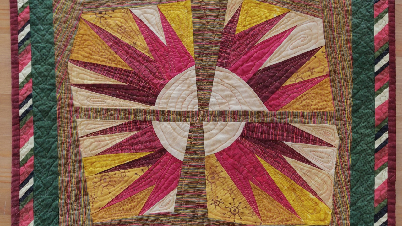 Winter Solstice Quilt’ by Karen Arends, featuring a white circular, spiraling sun at the center split into four quadrants, each with bold radiating points in burgundy, magenta, gold, and cream. The quilt is bordered by green panels and multicolored diagonal stripes, evoking warmth, light, and the return of the Sun