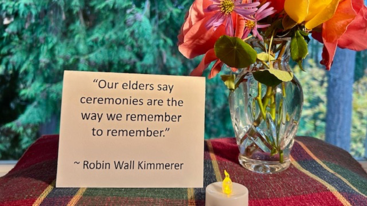 This close-up of a section of the Altar features a standing white card with a quote from Robin Wall Kimmerer that reads, “Our elders say ceremonies are the way we remember to remember.” Underneath the card is a colorful placemat and behind it is a blurred green outdoor background seen through a window.