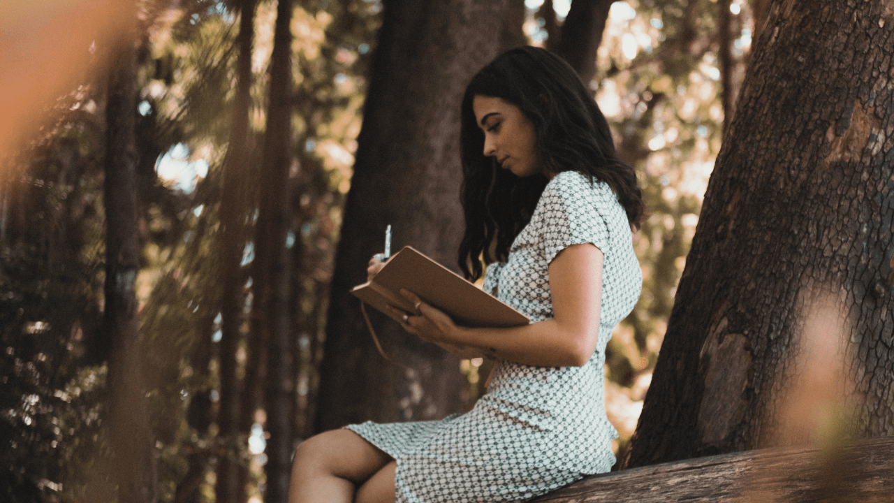 A woman sits on a trunk in the forest, writing and deep in thought. She has long dark hair and wears a cream-colored dress.