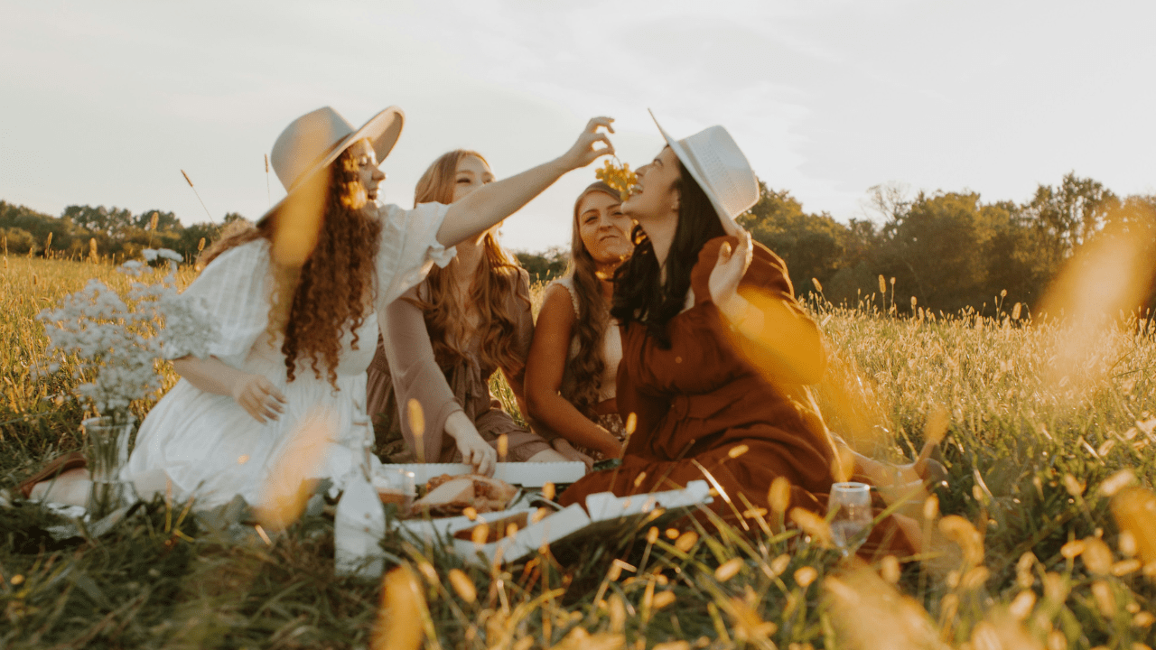 A group of four women is sitting outside in a field, chatting and enjoying some food. They appear very relaxed and happy.