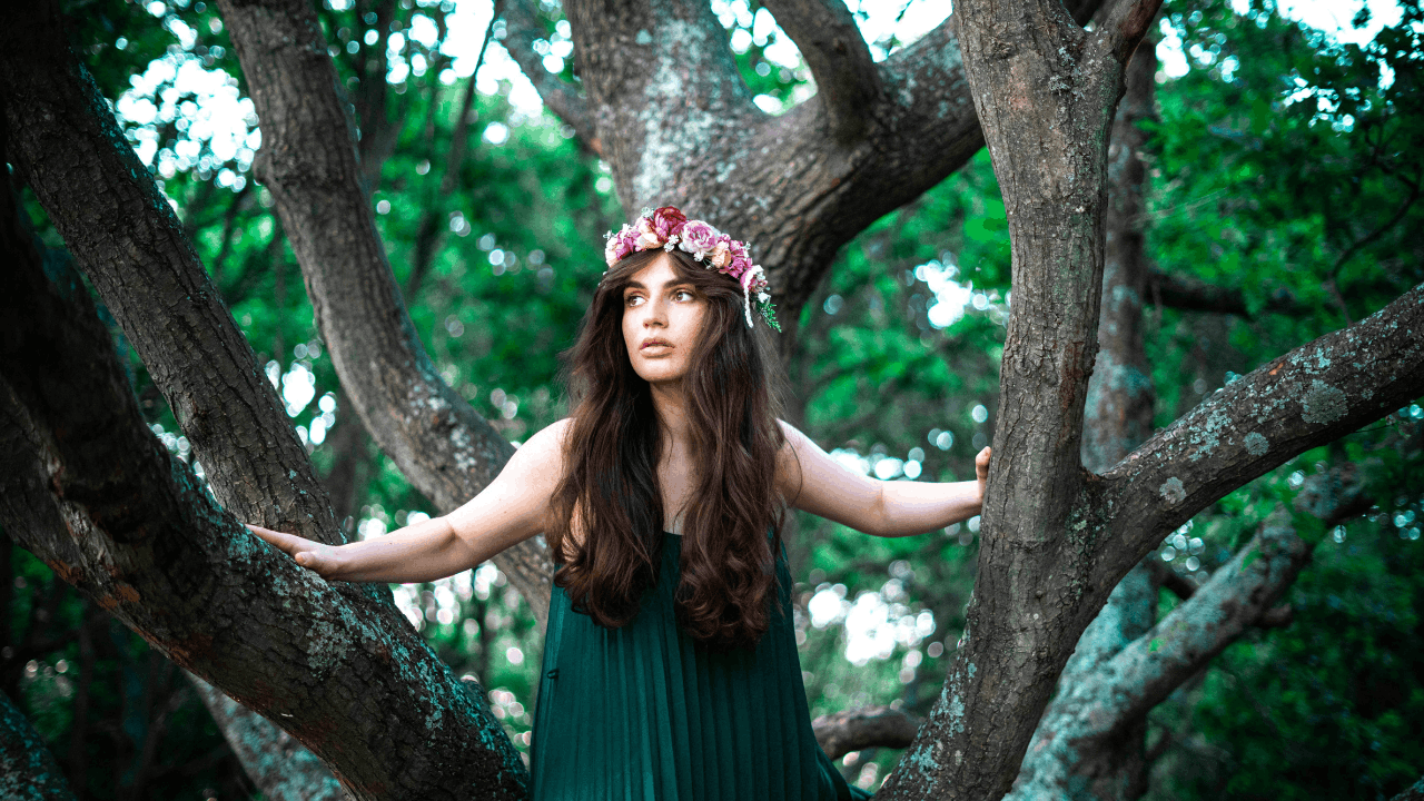 A woman is standing in a tree. She wears a long green dress and has flowers in her hair.
