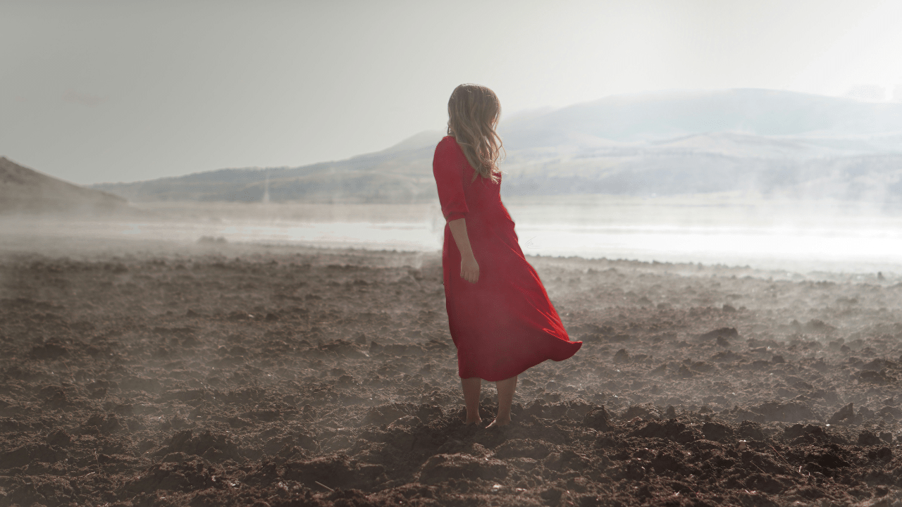 A woman is standing in the middle of a valley. She is wearing a long flowy red dress. The weather is very windy.
