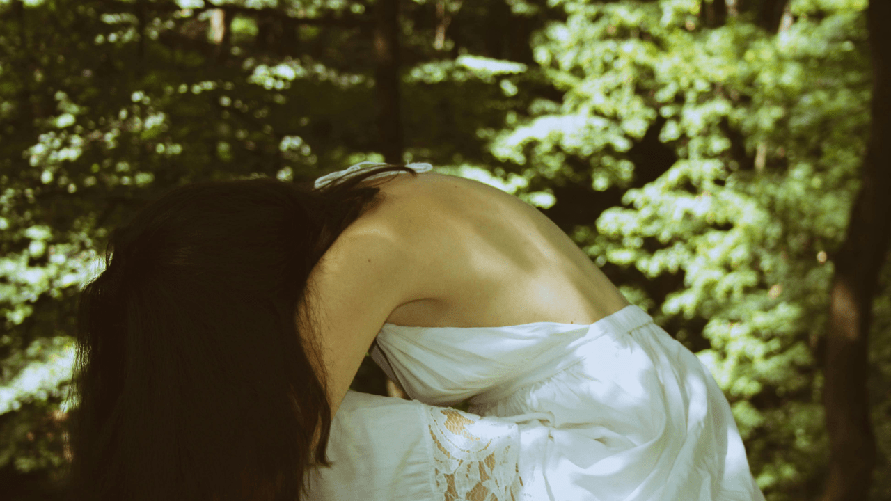 A woman is sitting in a forest. She is slumped forward, her face hidden by her long brown hair.