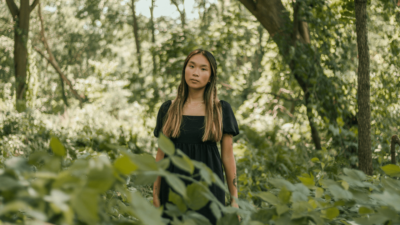 A woman stands alone in a beautiful forest, appearing serious and connected to the land.