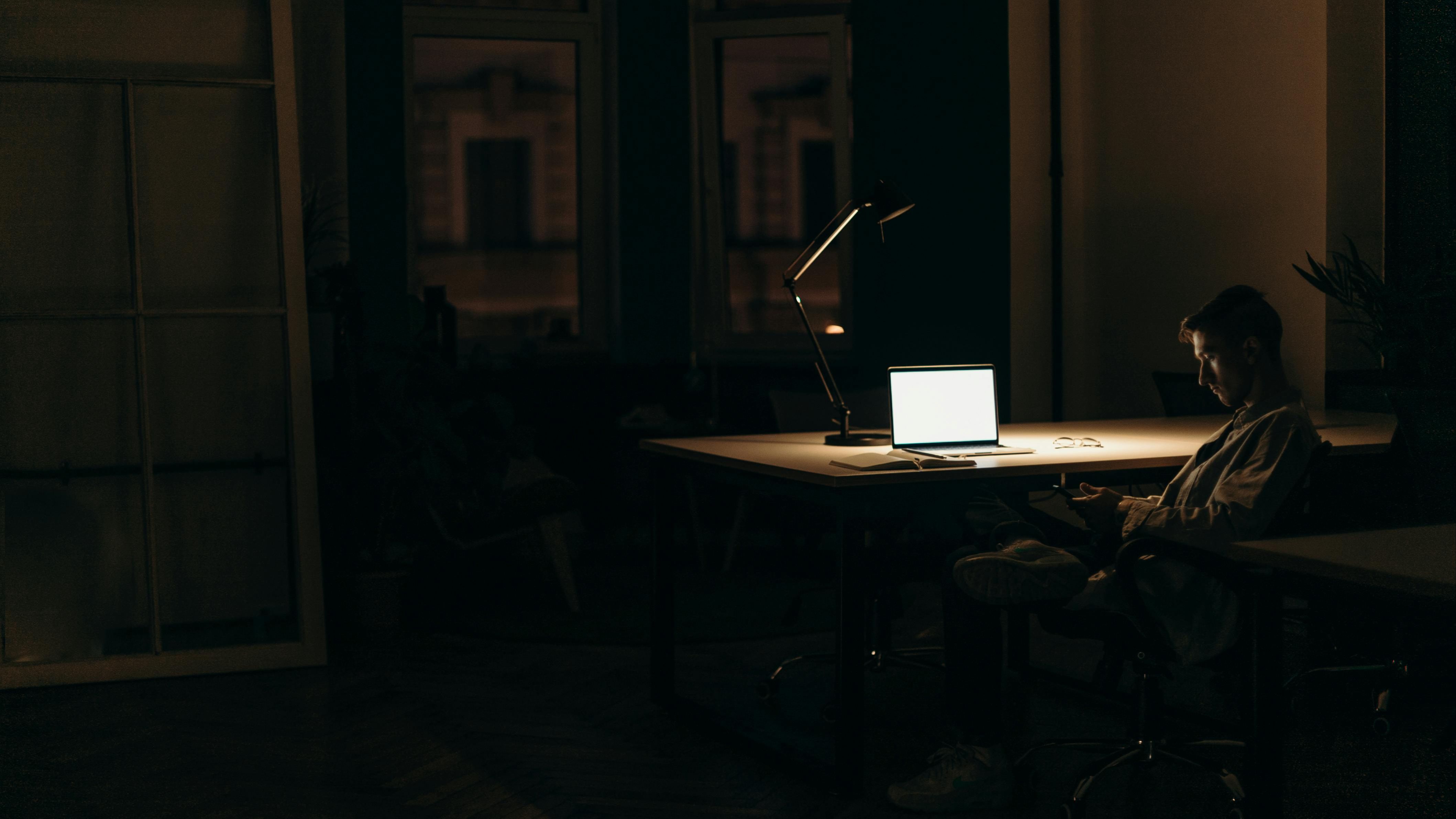 A man sitting at a desk lit with a single lamp, looking at his phone pensively, indicating his silence.