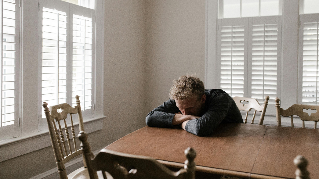 Man sitting at a table with his head on his arms, looking upset, next to an empty chair, reflecting feelings of self-doubt and emotional distance.