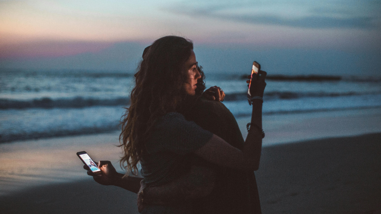 Married couple embracing at sunset on the beach while distracted by smartphones.