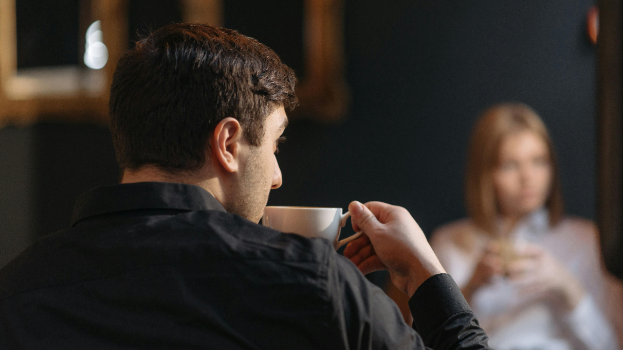 A man drinking a cup of coffee sits in front of a woman. Both look to the side, away from each other, seemingly disconnected and not listening to each other.
