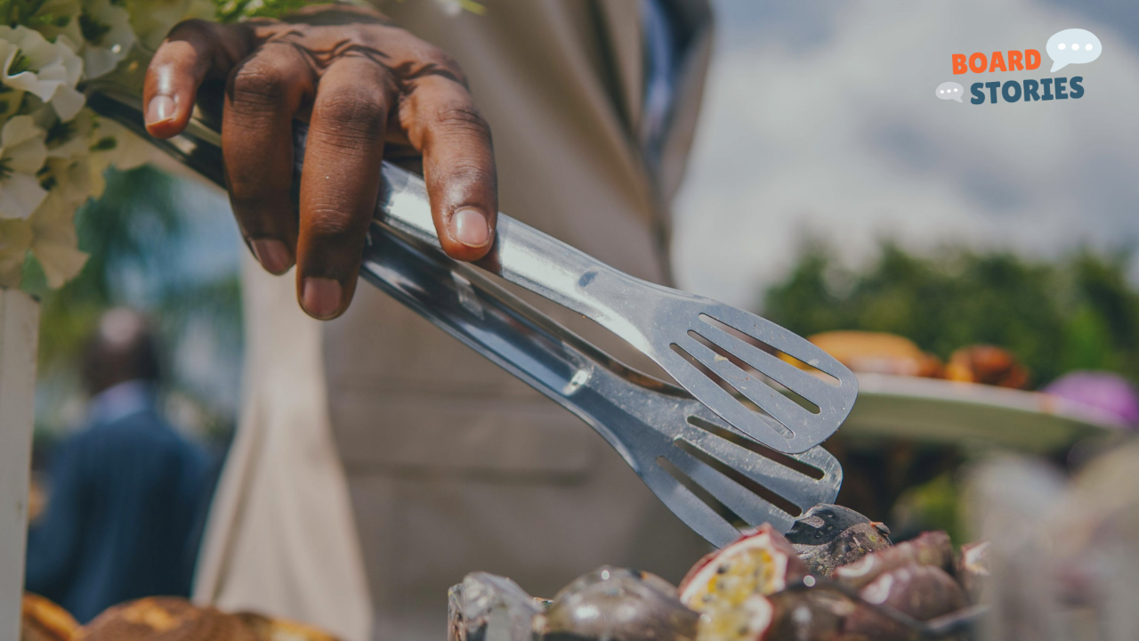 Close-up of a person using metal tongs to serve salad at an outdoor buffet table, with the “Board Stories” logo in the corner.