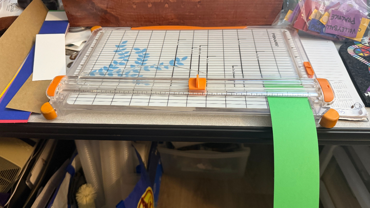 Cutting green construction paper into visual schedule cards using a Fiskars paper trimmer on a busy mom’s desk with craft supplies