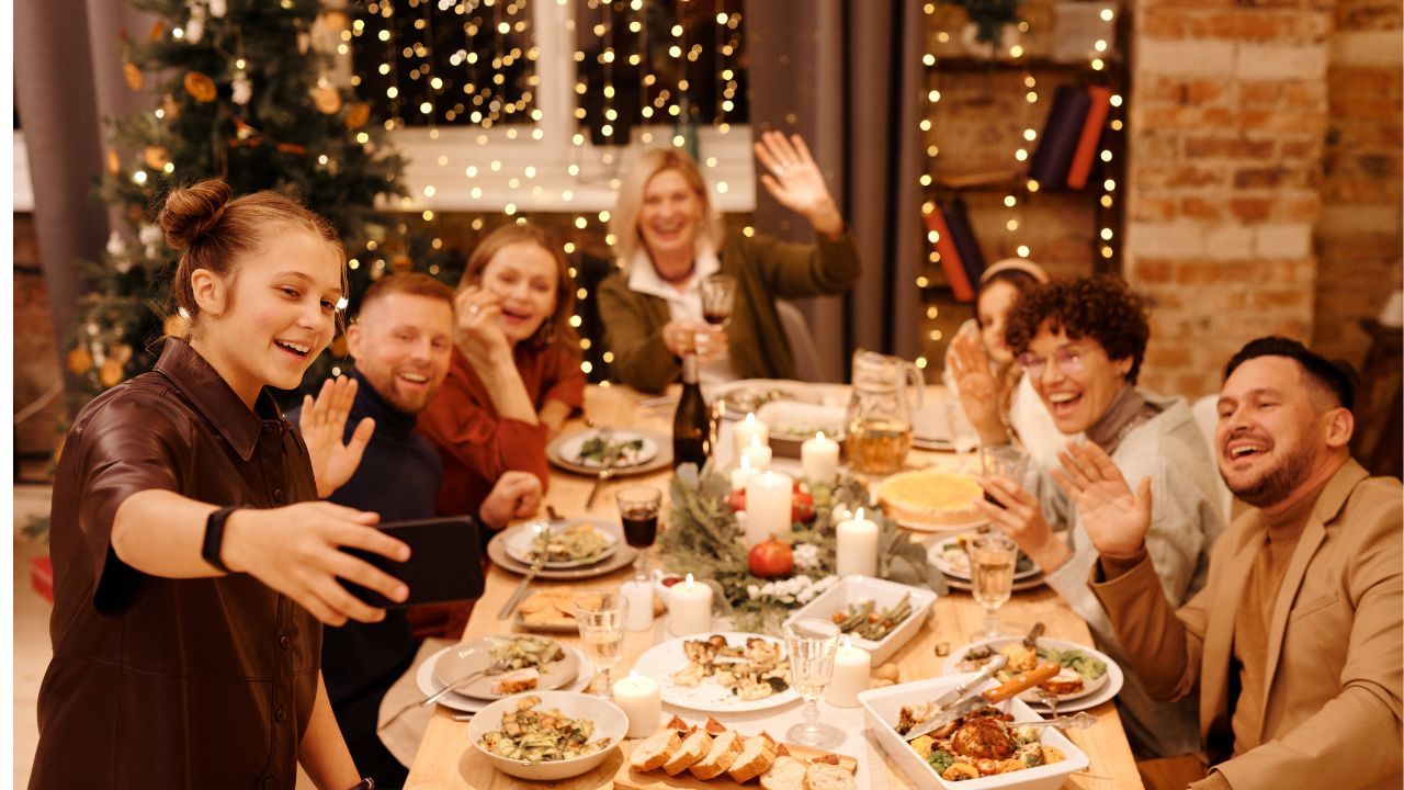 A joyful blended family gathered around a holiday dinner table, smiling and waving while taking a selfie, with twinkling lights and a Christmas tree in the background.