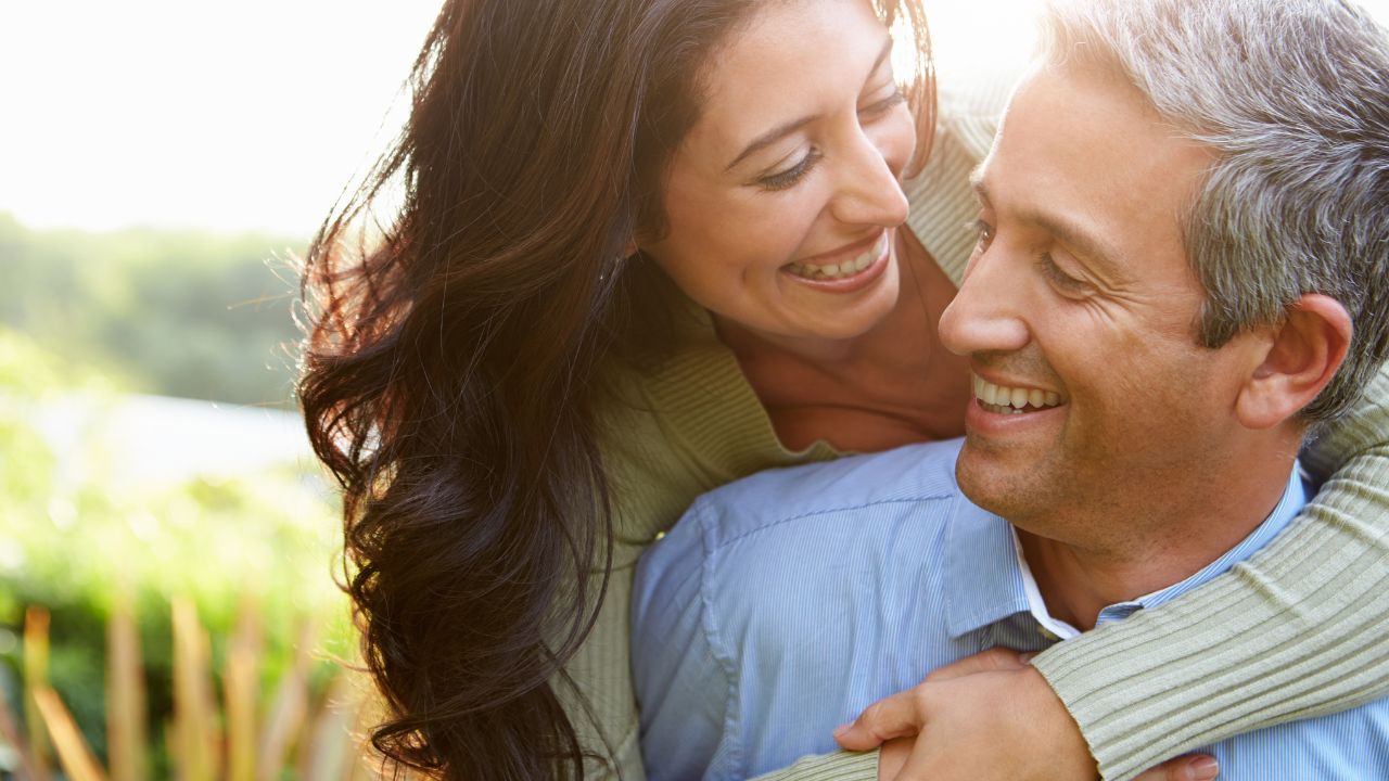 Middle-aged couple smiling outdoors as the woman hugs the man from behind, representing love and connection in midlife after divorce or loss.