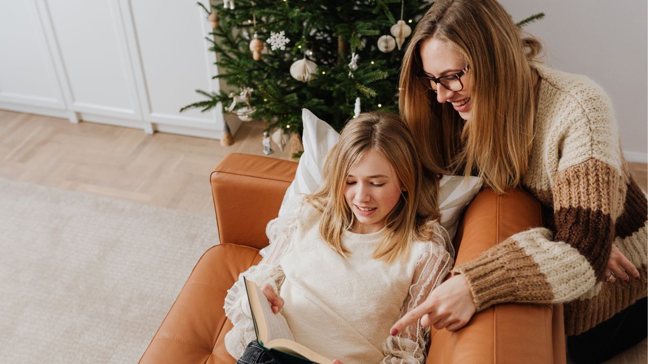 A smiling midlife mom and her young adult daughter sitting together on a couch near a decorated Christmas tree, looking at a book and enjoying relaxed holiday time together.