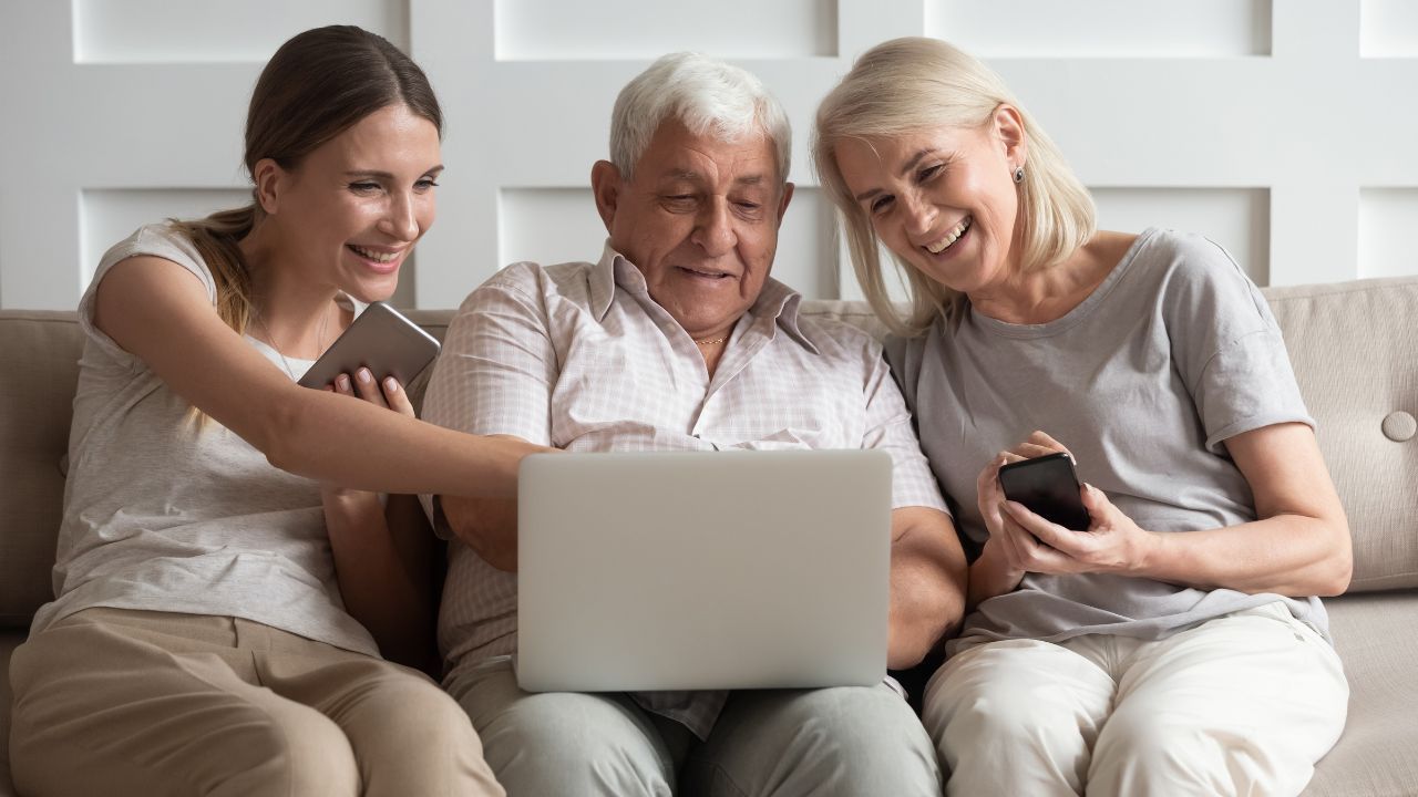 A midlife woman, an older man, and a young adult woman sit together on a couch smiling while looking at a laptop. The scene captures multigenerational connection and the joy of being present during the holidays.